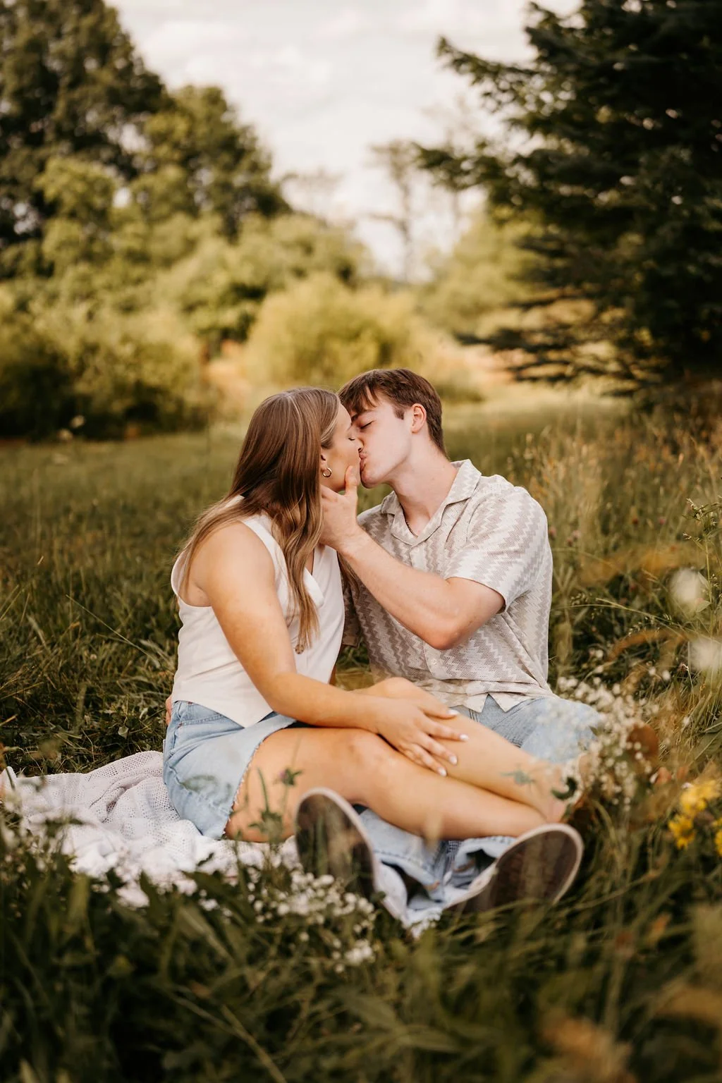 A young couple is sitting on a blanket in a grassy field, kissing with a river and green trees in the background during daytime.