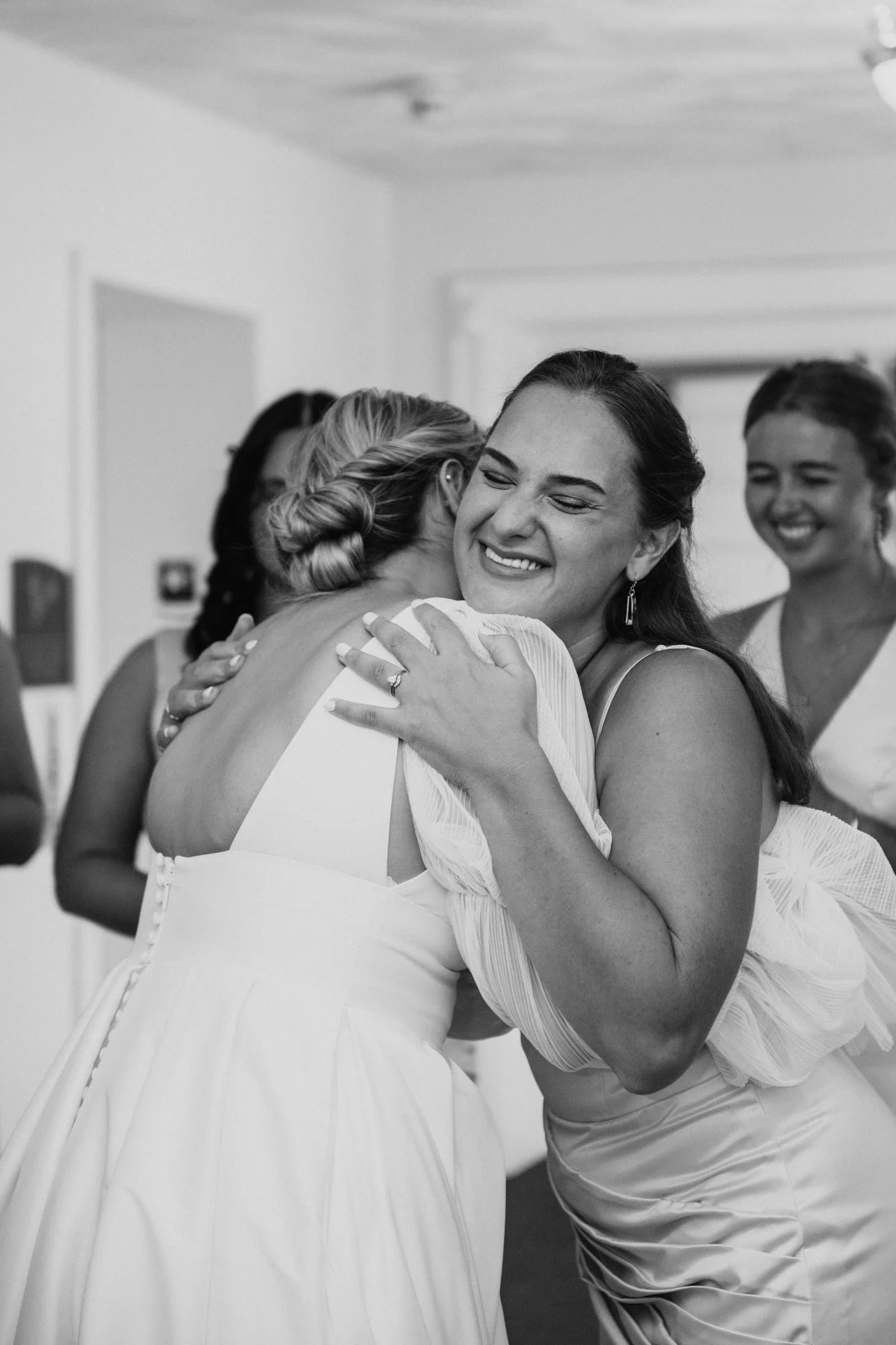 A woman hugging another woman while smiling, with two other women smiling in the background, in a wedding setting.