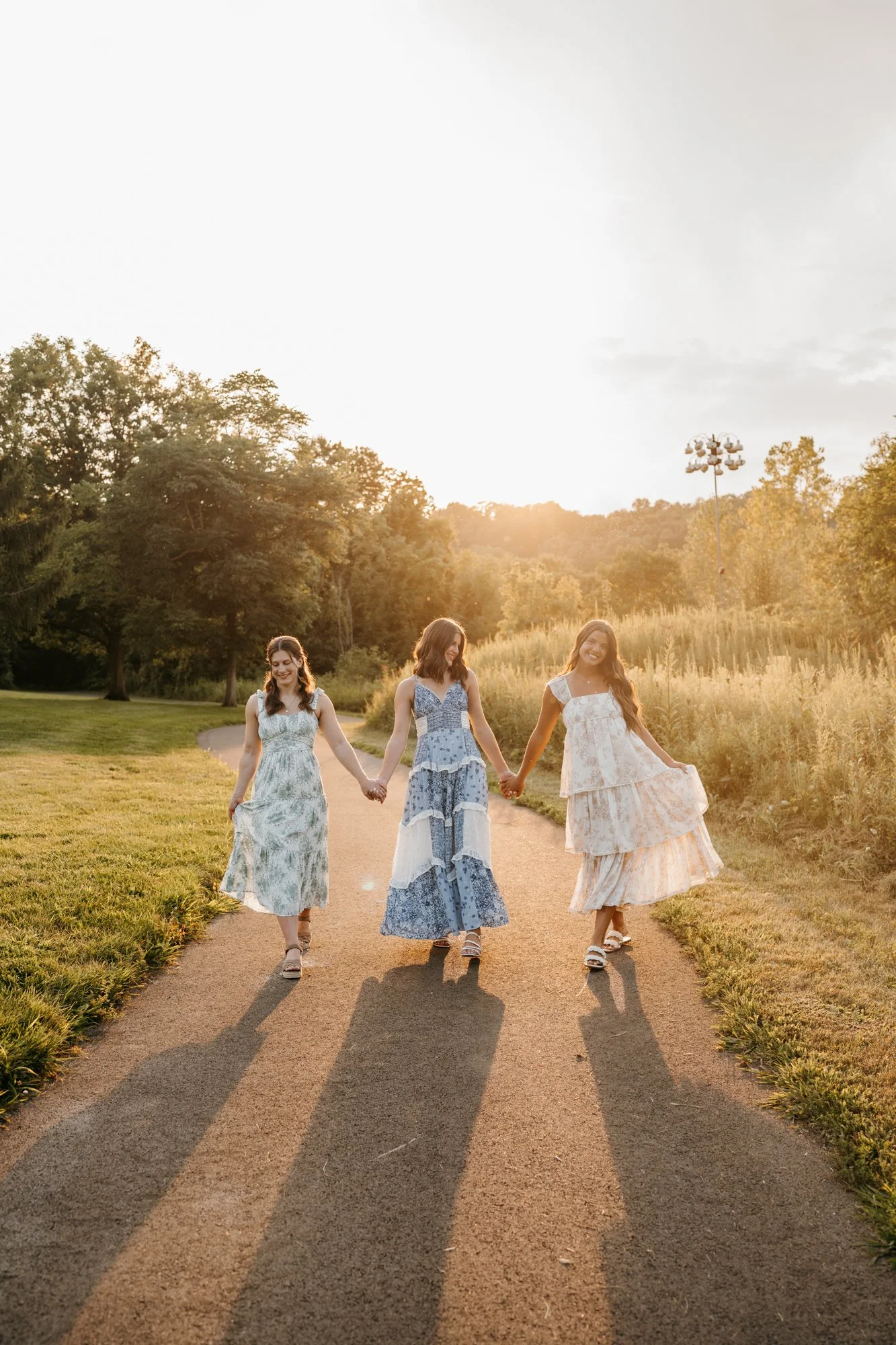 Three women walking hand in hand on a path in a park during sunset, wearing long flowy dresses.