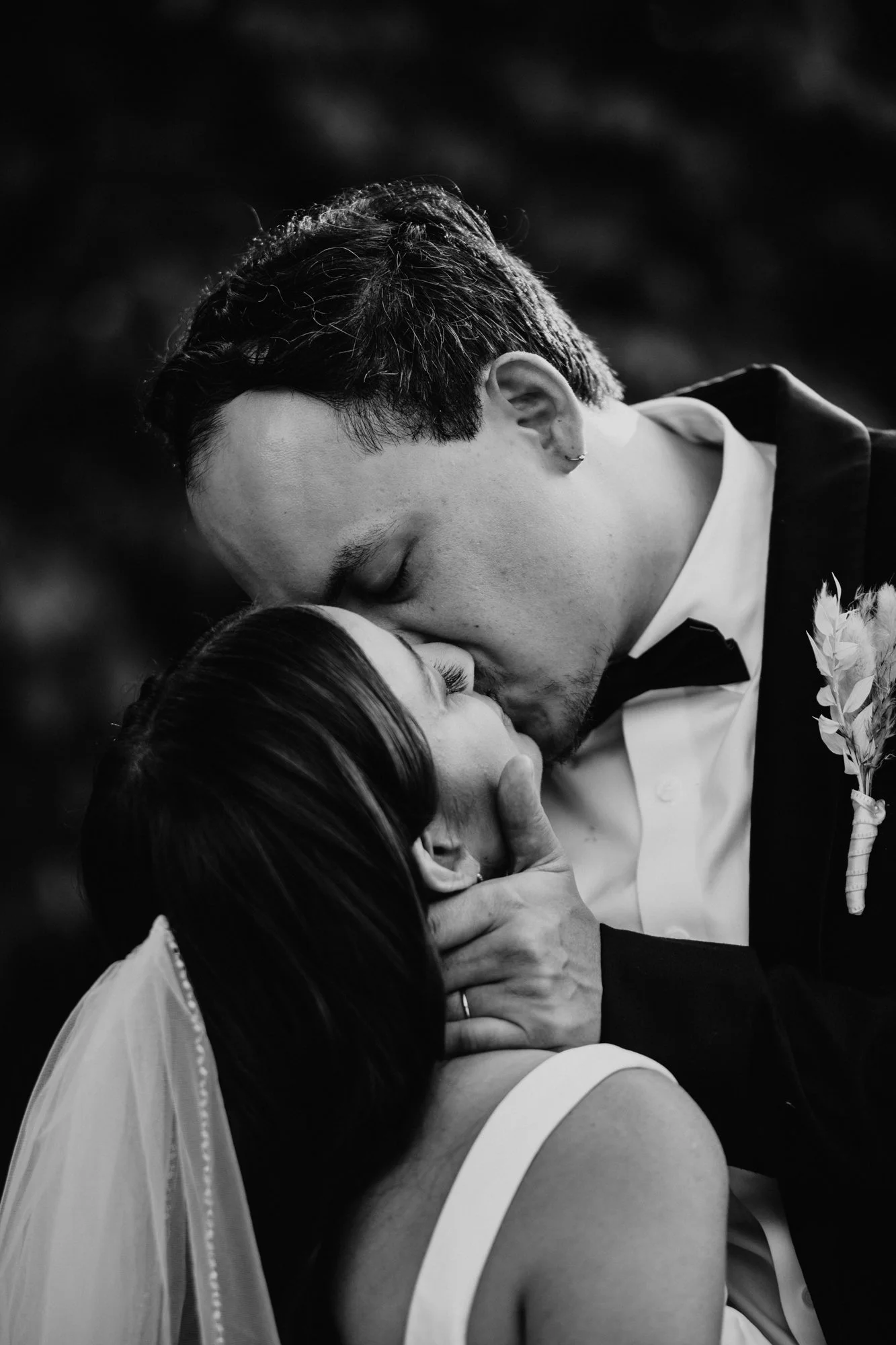 Black and white photo of a couple kissing at their wedding, with the groom gently holding the bride's face.
