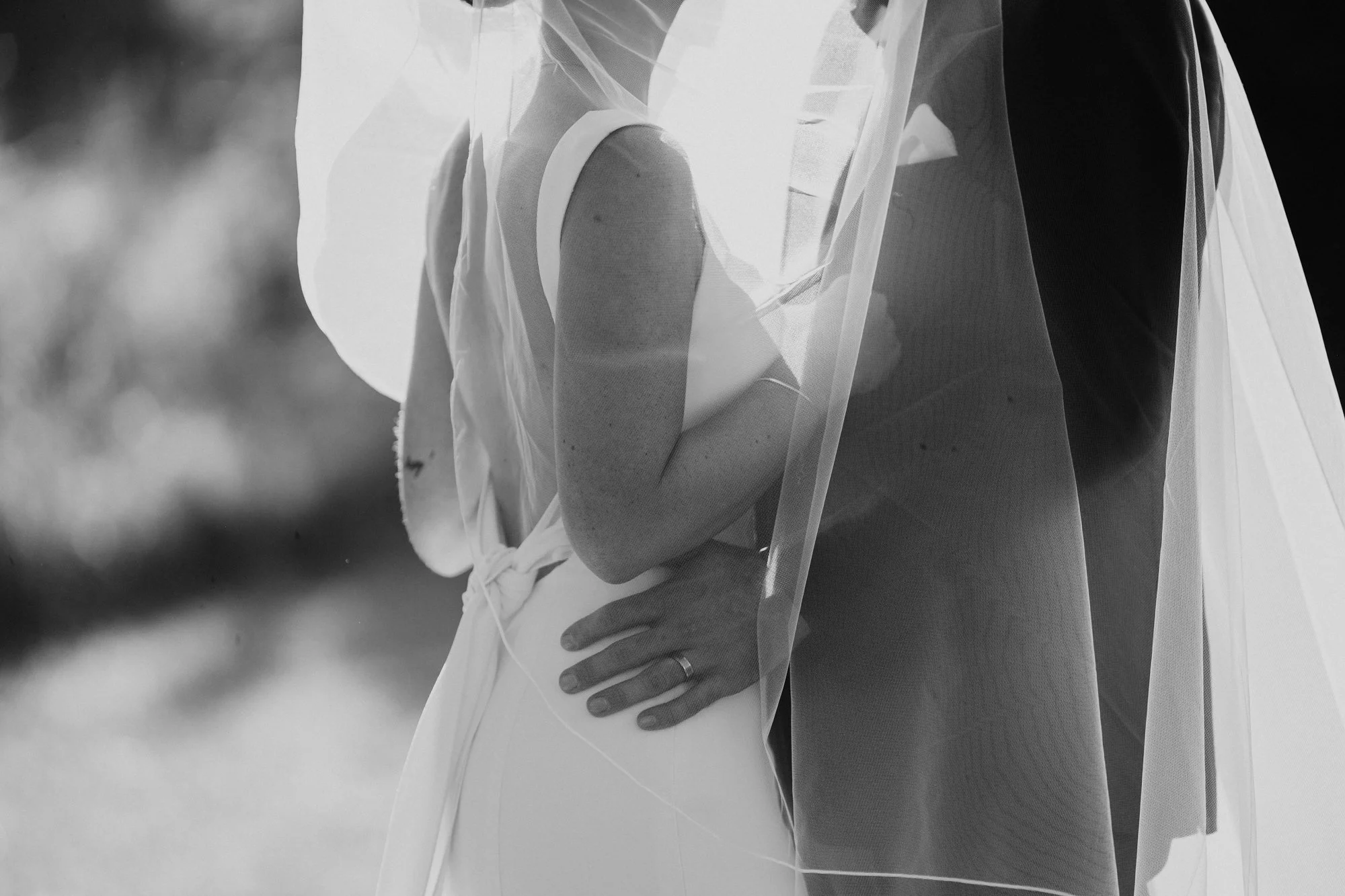 Close-up of a bride and groom holding hands, with the bride's hand resting on her hip and the groom's hand on her waist, both dressed in wedding attire, the bride wearing a veil and the groom in a suit, during a wedding ceremony.