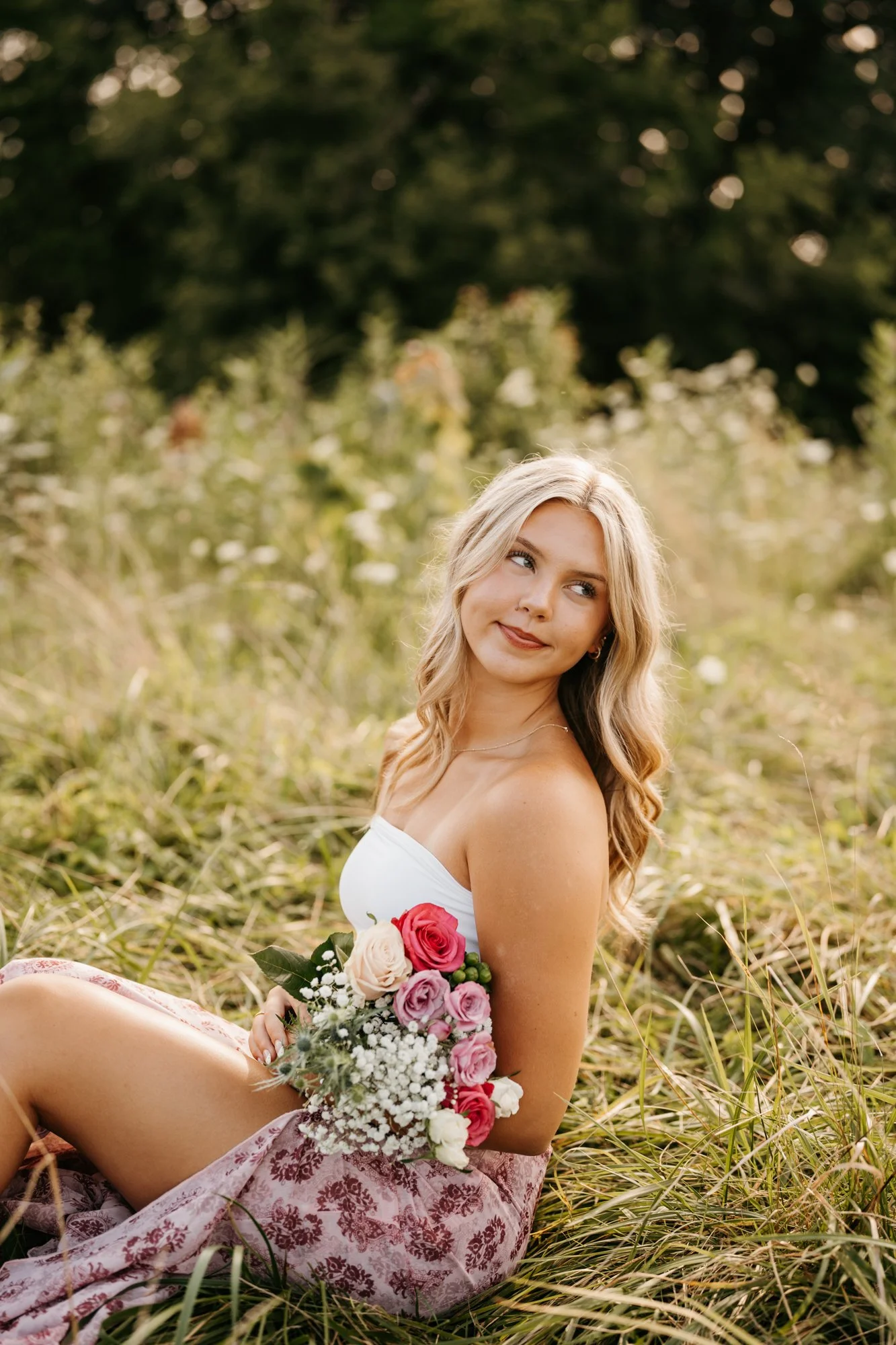 A young woman with long blonde hair sitting outdoors on grass, holding a bouquet of pink, white, and red roses, with a peaceful expression, in a natural setting.