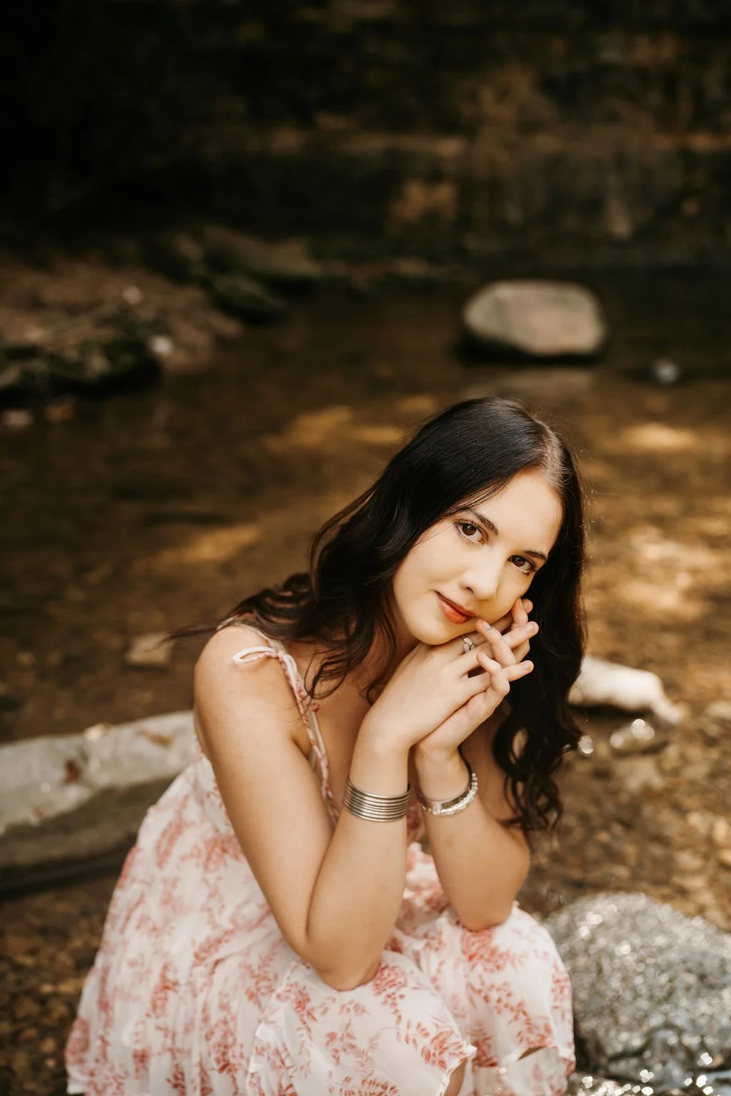 A woman with long dark hair smiling and resting her chin on her hands, sitting by a shallow creek with rocks in the background.