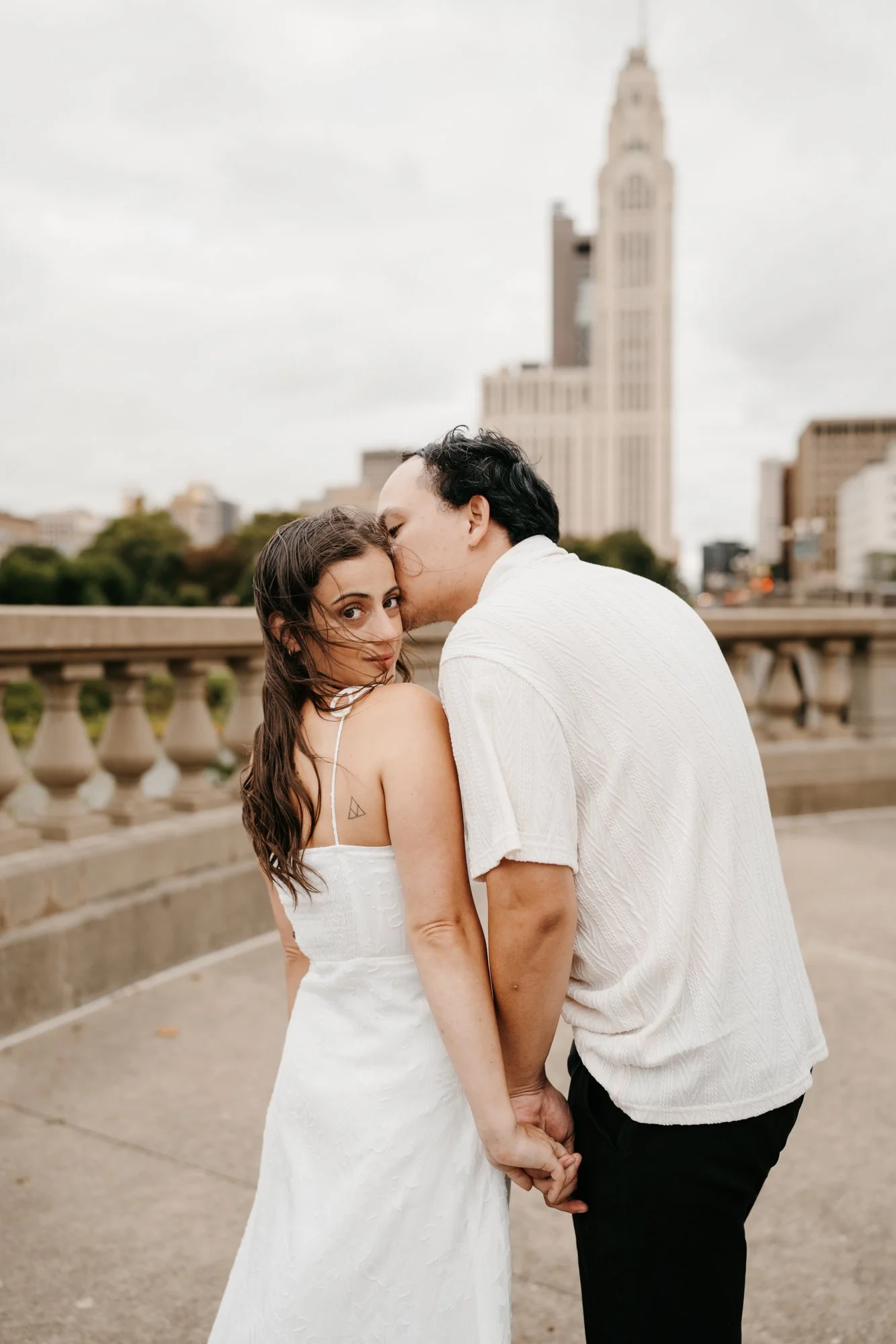 A couple holding hands on a bridge in an urban setting, with a city skyline and tall building in the background. The woman is looking at the camera, and the man is kissing her temple.