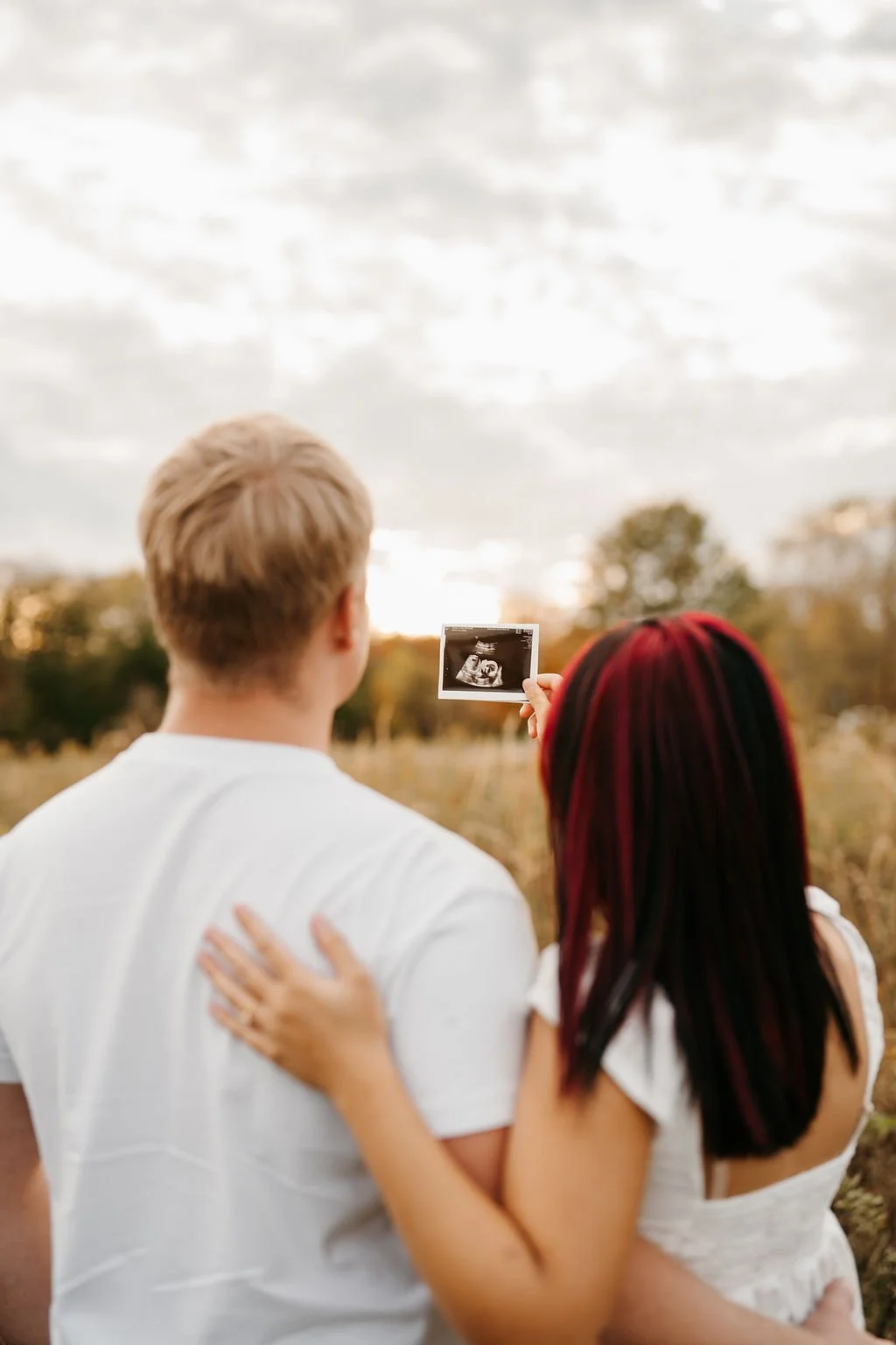 A couple stands in a field during sunset, with the woman holding an ultrasound photo, both with their backs to the camera.