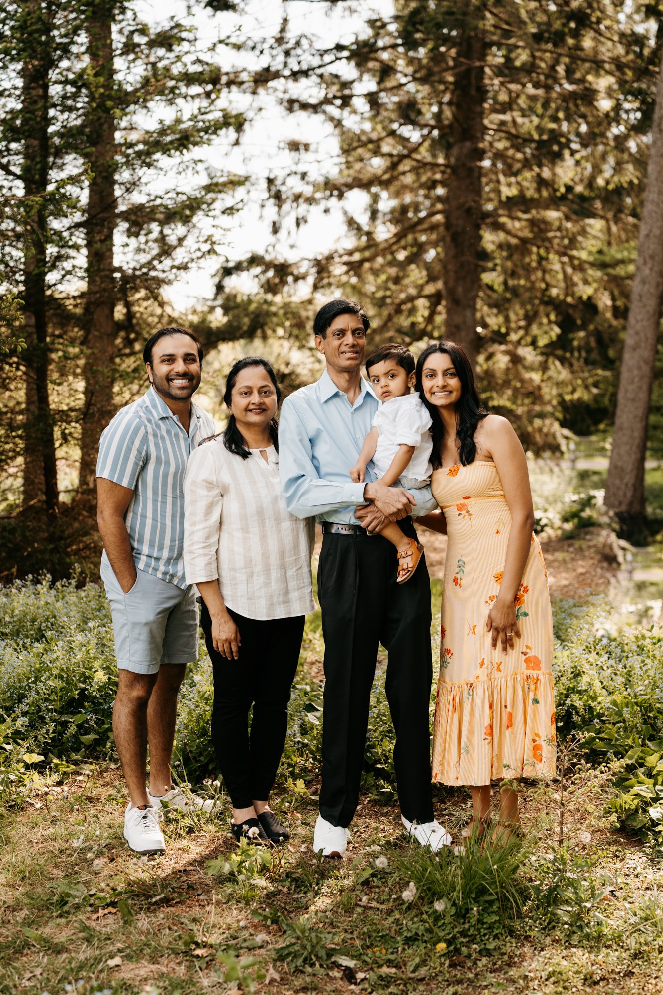 Group of five people, including a young child, family posing outdoors in a forested area with tall trees and greenery.