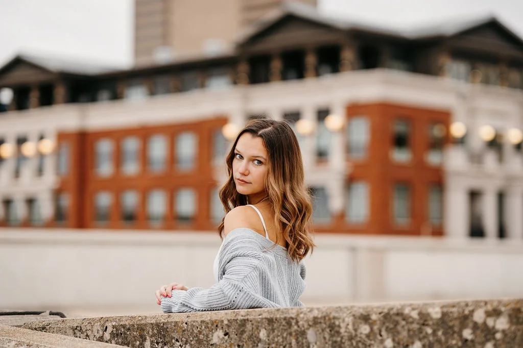 Young woman with wavy brown hair looking at the camera, standing outdoors with a cityscape behind her, wearing a gray off-shoulder sweater.