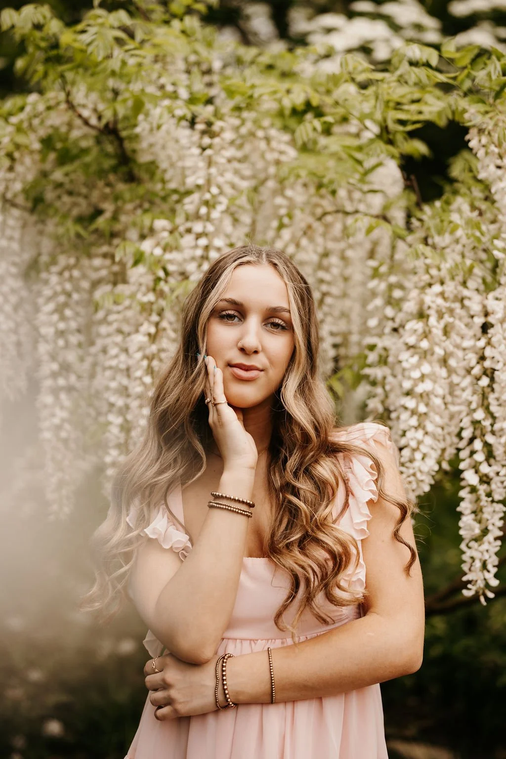 A young woman with long, wavy brown hair, wearing a light pink dress with ruffled sleeves, stands in front of a background of white flowering trees. She is touching her face gently with her right hand, wearing bracelets on both wrists, and has a calm