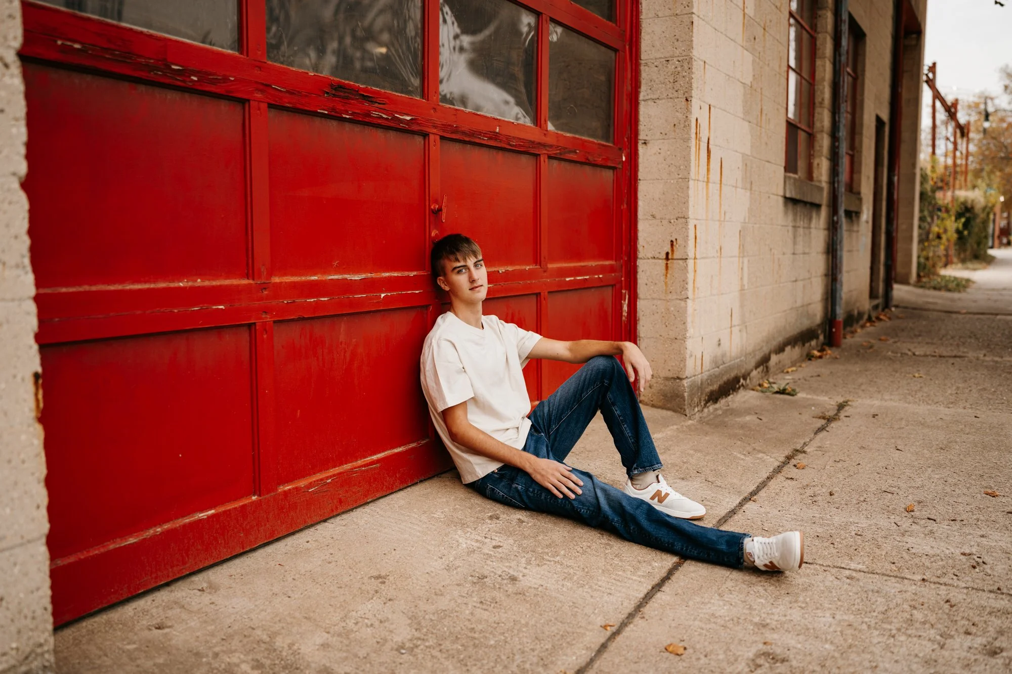 A young person sitting on the sidewalk leaning against a large red industrial door with a beige brick wall to the right, in an urban setting.