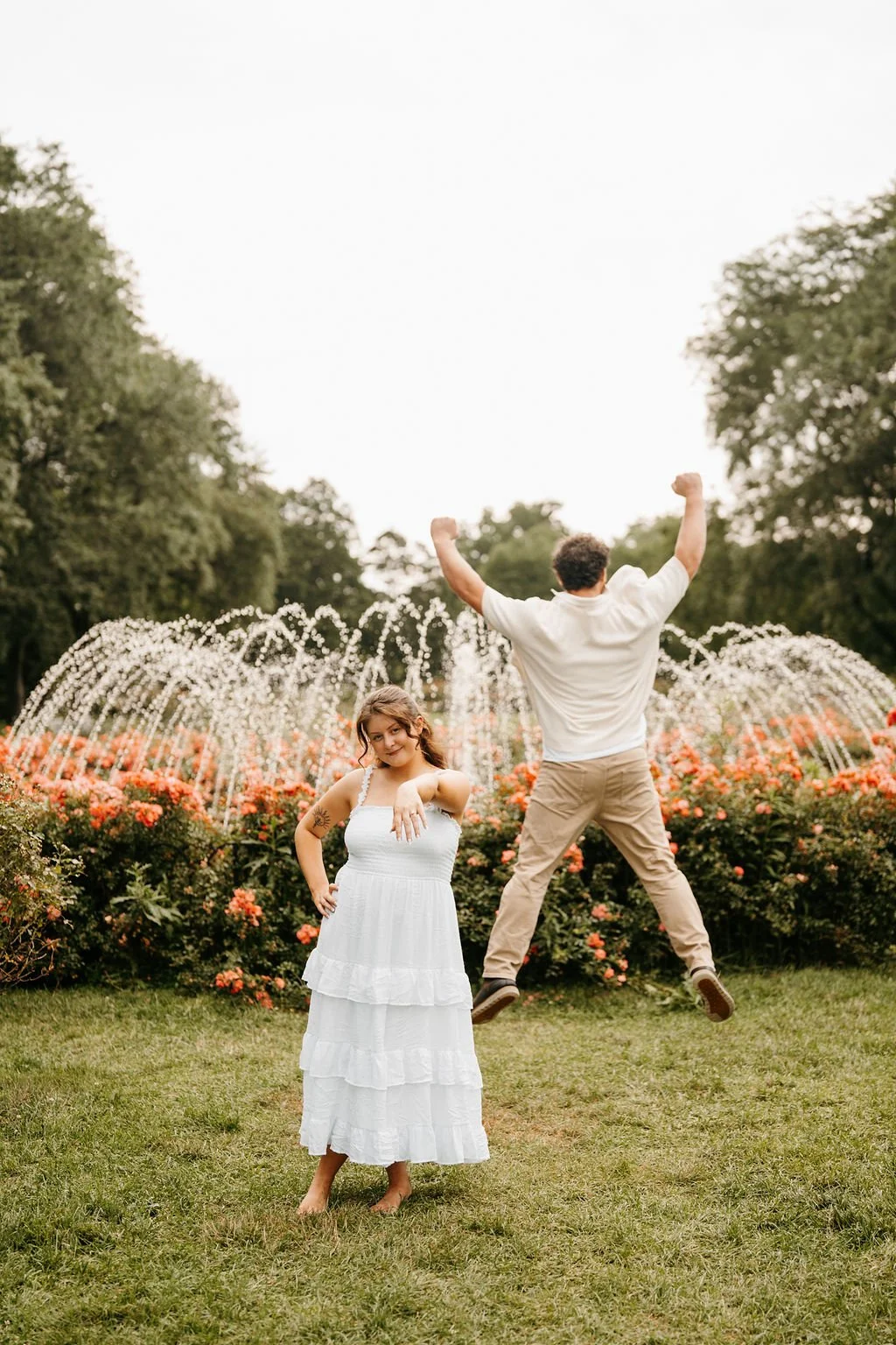 A woman in a white dress standing on grass with a tattoo on her arm, and a man in khaki pants jumping with arms raised in front of a fountain with water jets and pink flowers, surrounded by green trees.