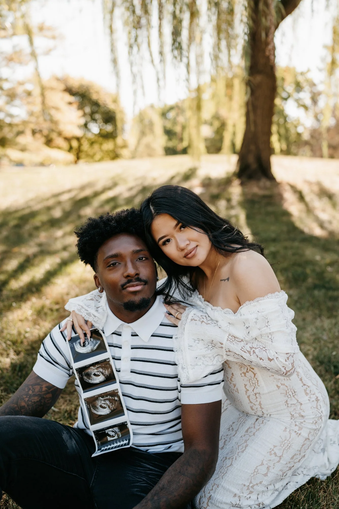 A man and woman sitting on grass outdoors, with the woman resting her head on the man's shoulder. The man has curly hair and tattoos, holding ultrasound images, while the woman with long black hair has a small tattoo on her chest and wears a white la