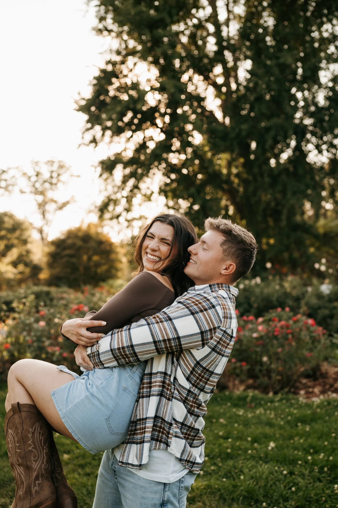 A smiling woman being carried by a man in a park during sunset, with trees and blooming flowers in the background.