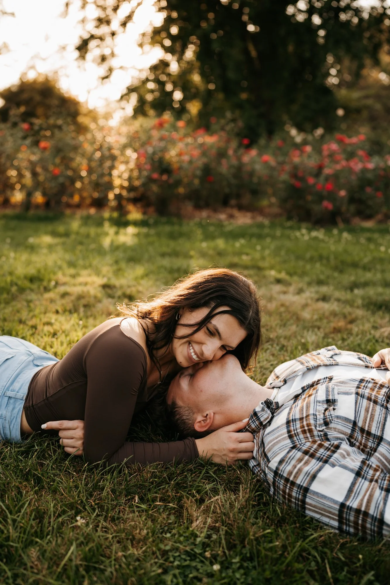 A young couple lies on the grass outdoors during sunset. The woman is smiling and leaning over the man, who is lying on his back. They share a loving moment in a park with trees and flowering bushes in the background.