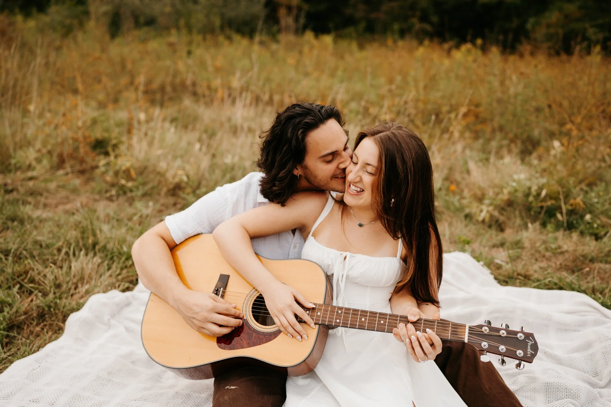 A young couple sitting on a white blanket in a field, smiling and playing guitar together during sunset.