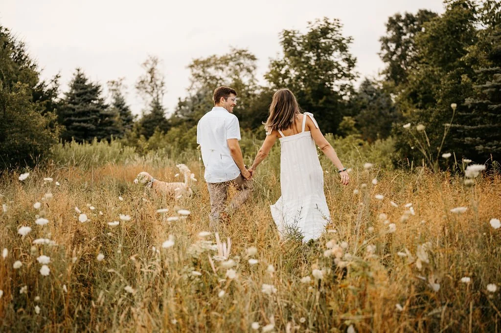 A couple holding hands walking through a grassy field with wildflowers, with a dog nearby, surrounded by trees and a cloudy sky.