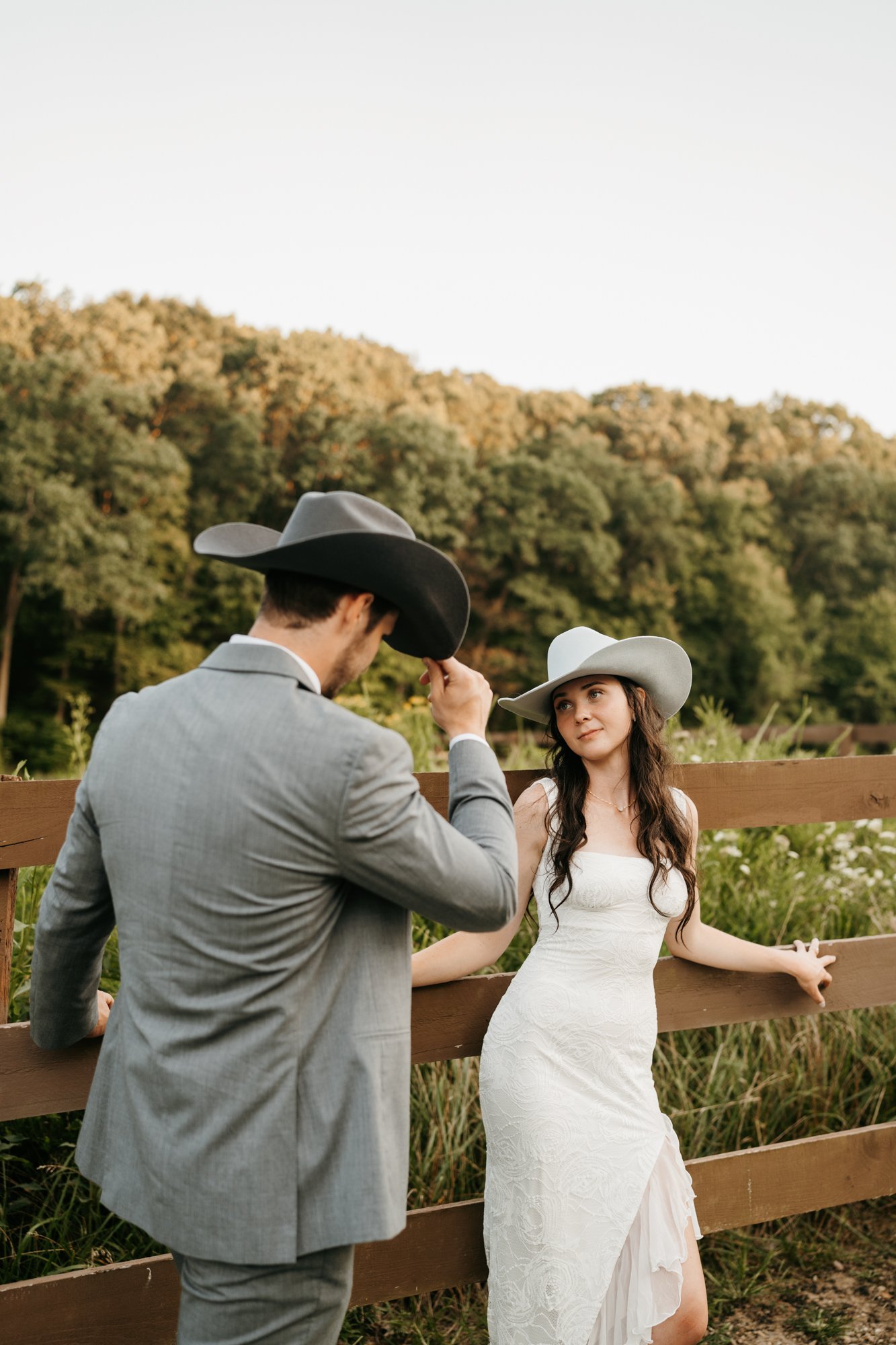 A woman in a white dress standing by a wooden fence with a man in a gray suit and cowboy hat, outdoors in a natural setting with trees and hills in the background.