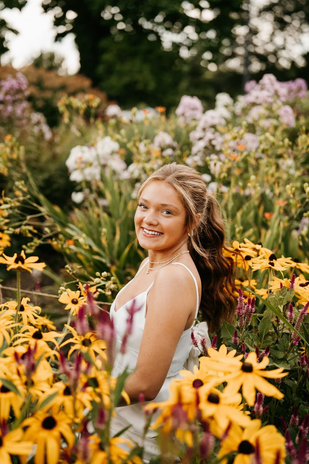 A young woman with long, wavy brown hair wearing a white dress and layered necklaces smiling while standing in a colorful flower garden.