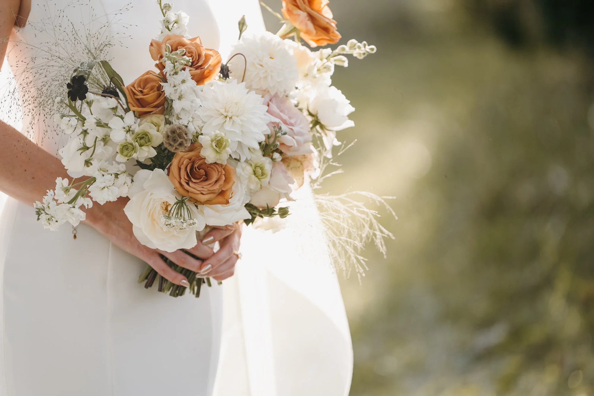Close-up of a bride holding a bouquet of flowers, including white, peach, and light pink blossoms, with blurred green background.