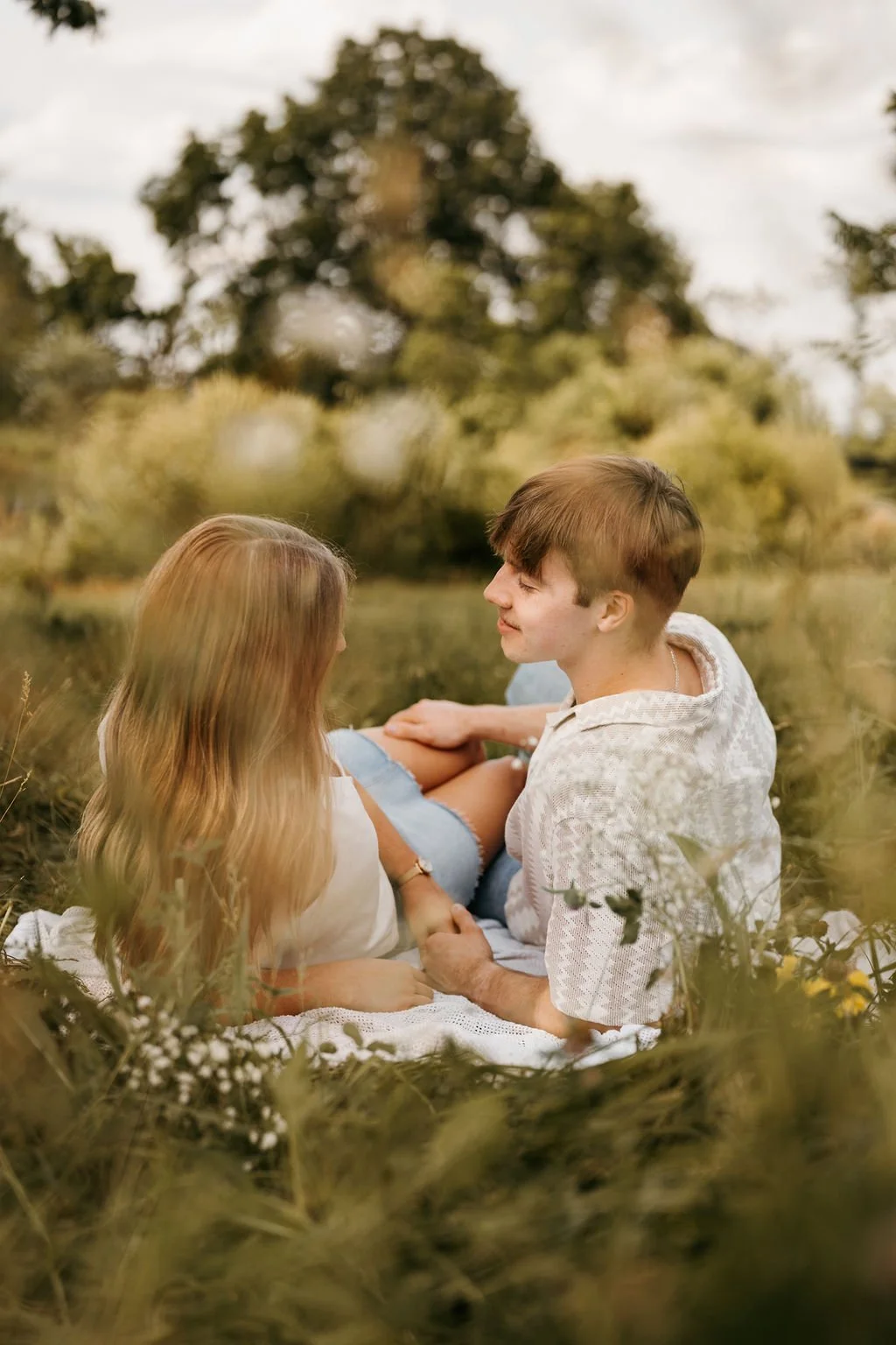 A couple sitting on a blanket in a grassy field, looking at each other and holding hands, with trees and a cloudy sky in the background.