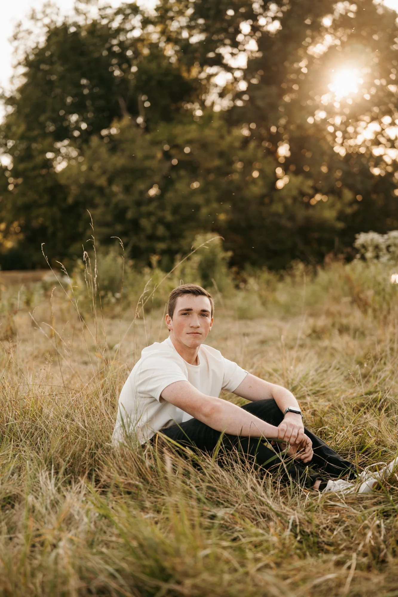 A young man sitting in a field of tall grass with trees and sunlight in the background during sunset.