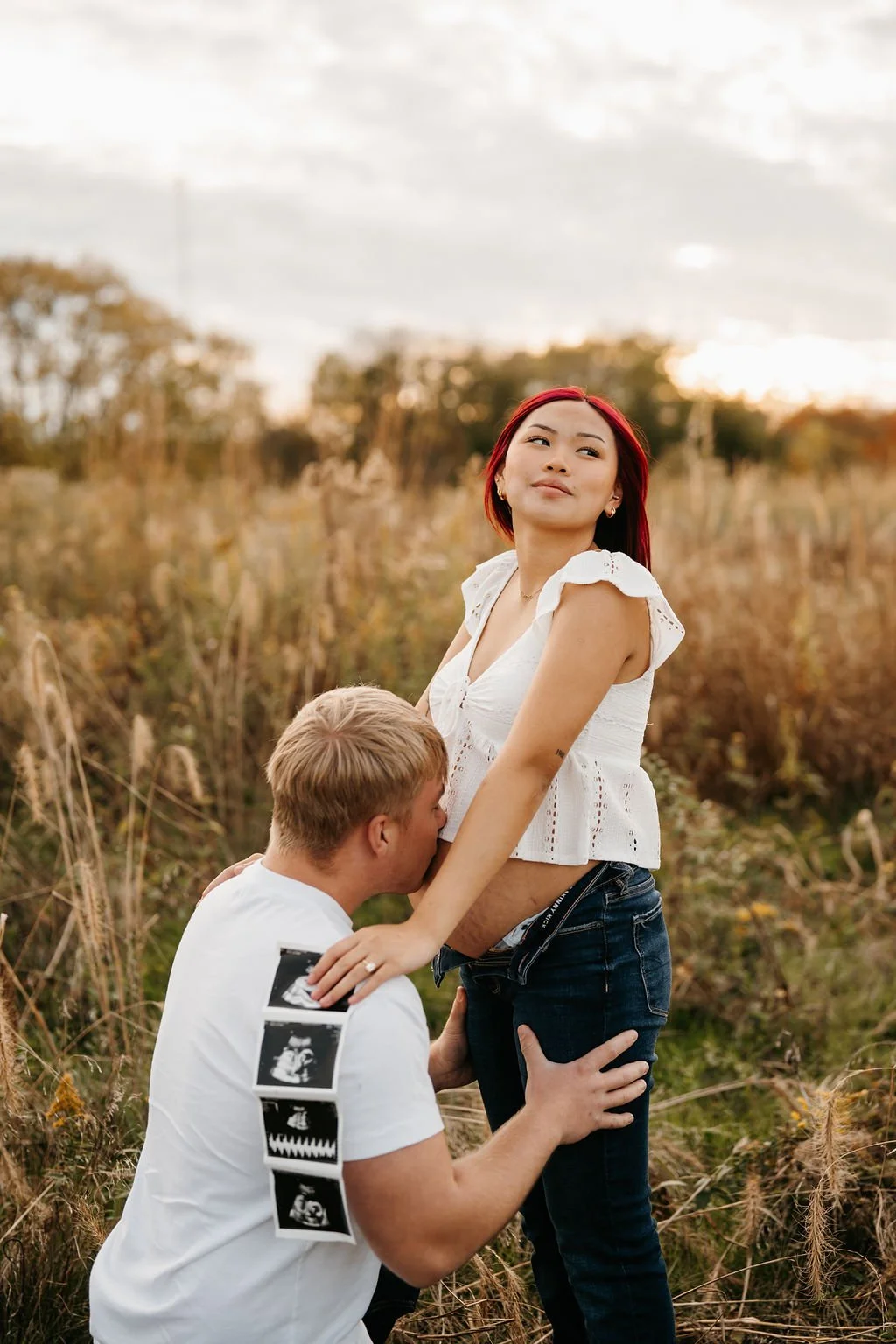 A woman with red hair and a white top standing outdoors in a field, with a man kneeling and giving her a kiss on her stomach, holding ultrasound pictures.