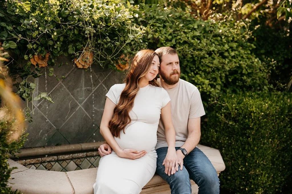 A pregnant woman with long brown hair in a white dress sitting on a garden bench with a man with a beard in a beige t-shirt in a lush outdoor setting.