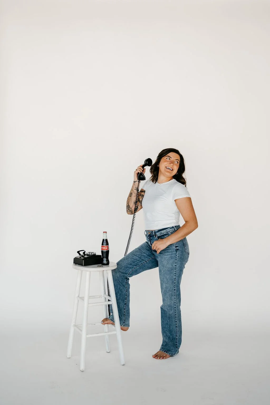 A woman with dark hair, tattoos, and wearing a white t-shirt and jeans, holding a vintage black rotary phone to her ear, while standing barefoot next to a white stool with a bottle of Coca-Cola and a vintage black camera on it, in a plain white studi