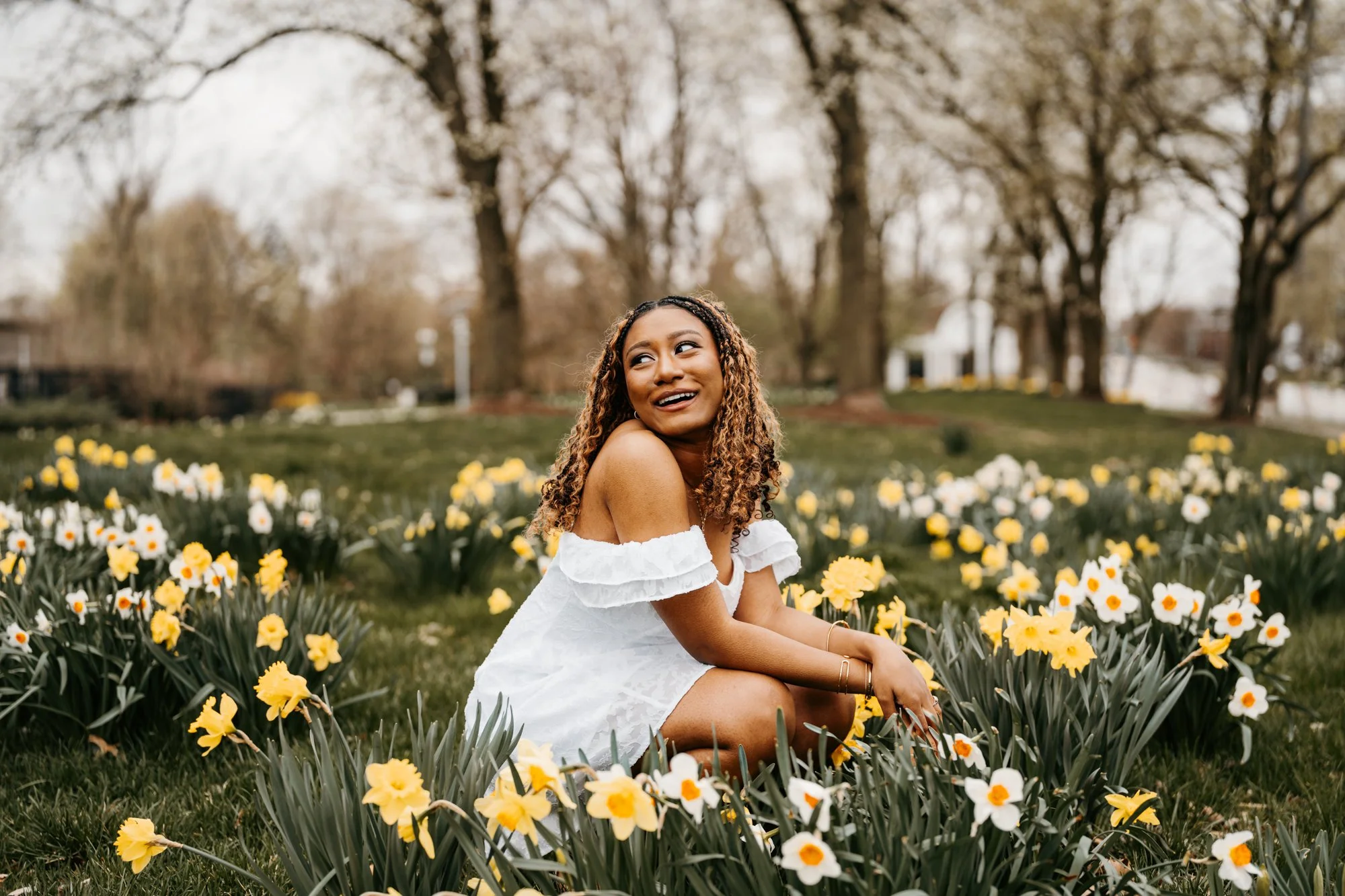 A woman with curly hair, wearing a white off-shoulder dress, is sitting on the grass surrounded by yellow and white flowers in a park on a spring day.
