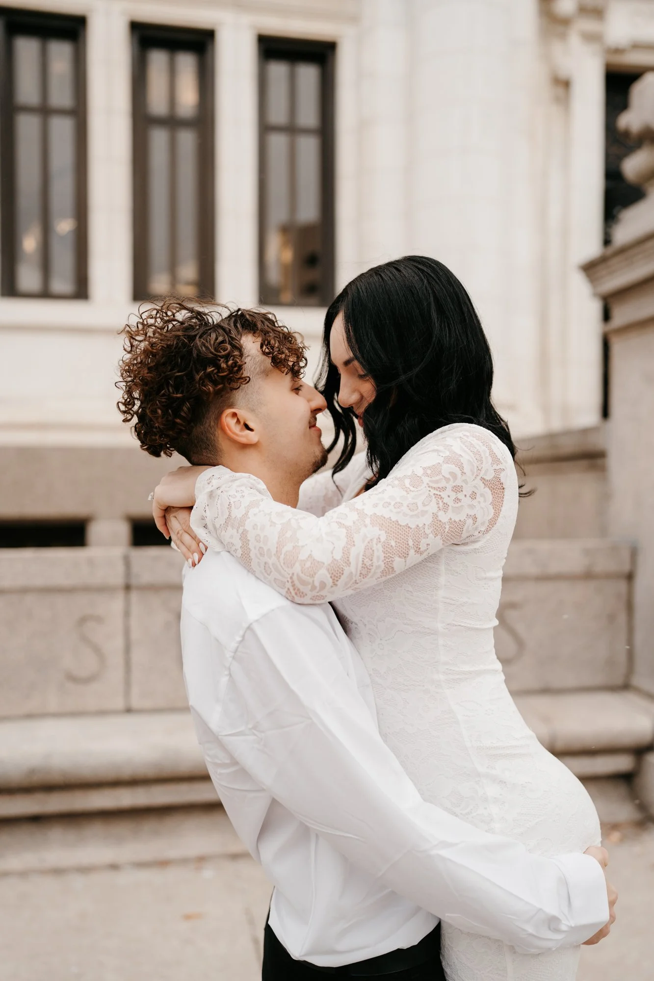 A couple embracing affectionately outside a building, with a man lifting a woman in a white lace dress.