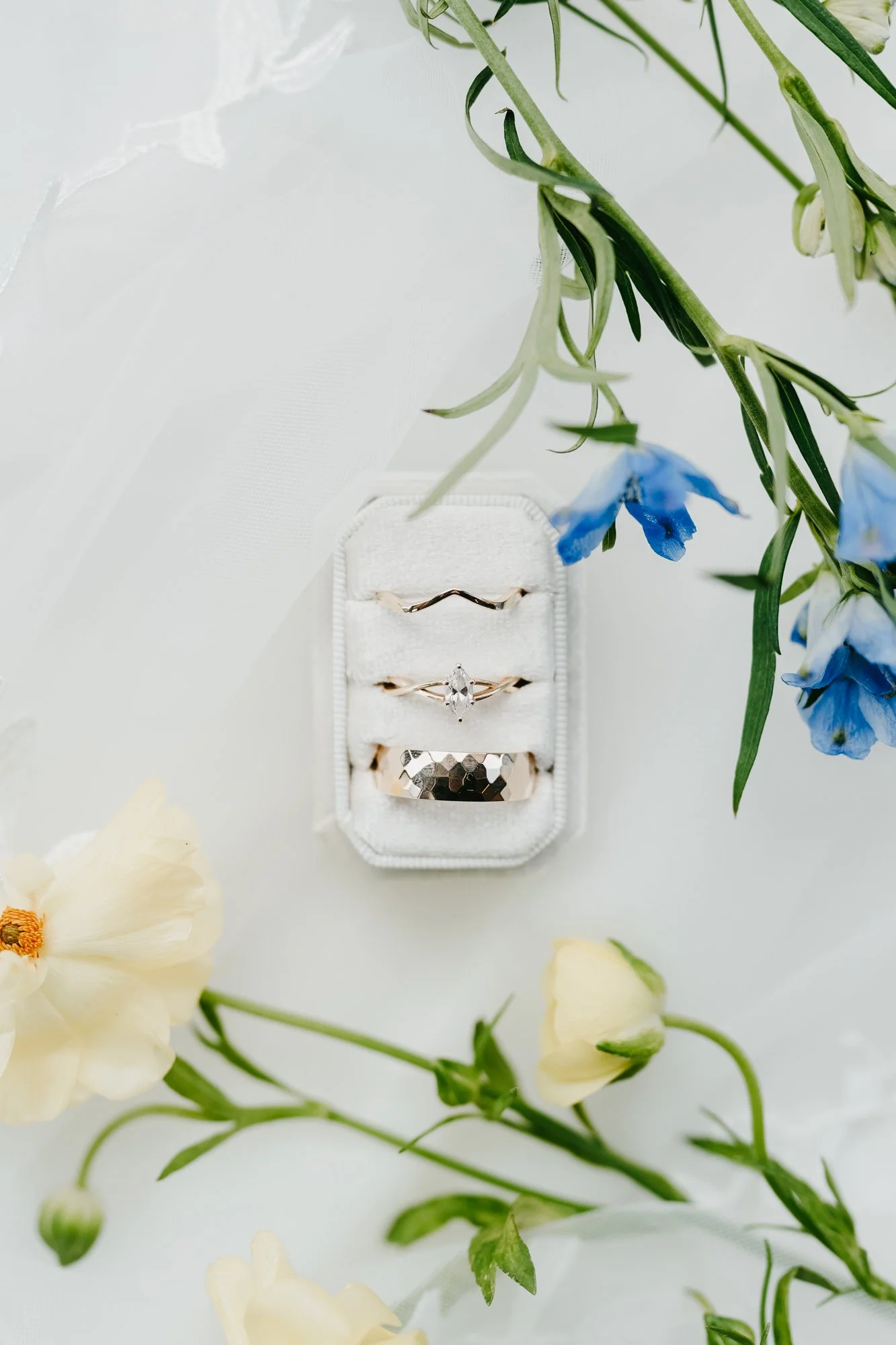 A small white ring box containing three rings with a floral arrangement of yellow and blue flowers surrounding it.