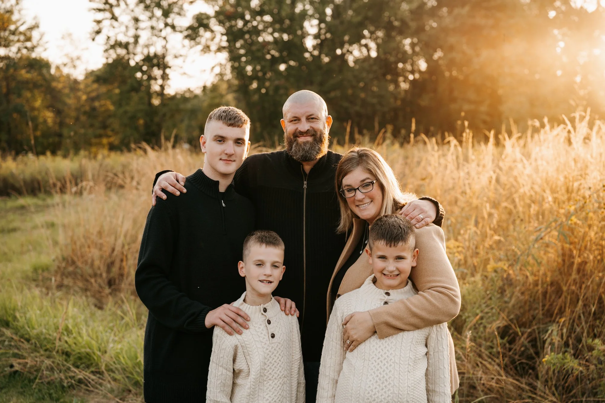 A family of six, including two children and two adults, standing outdoors in a field during golden hour sunset, smiling and embracing each other.