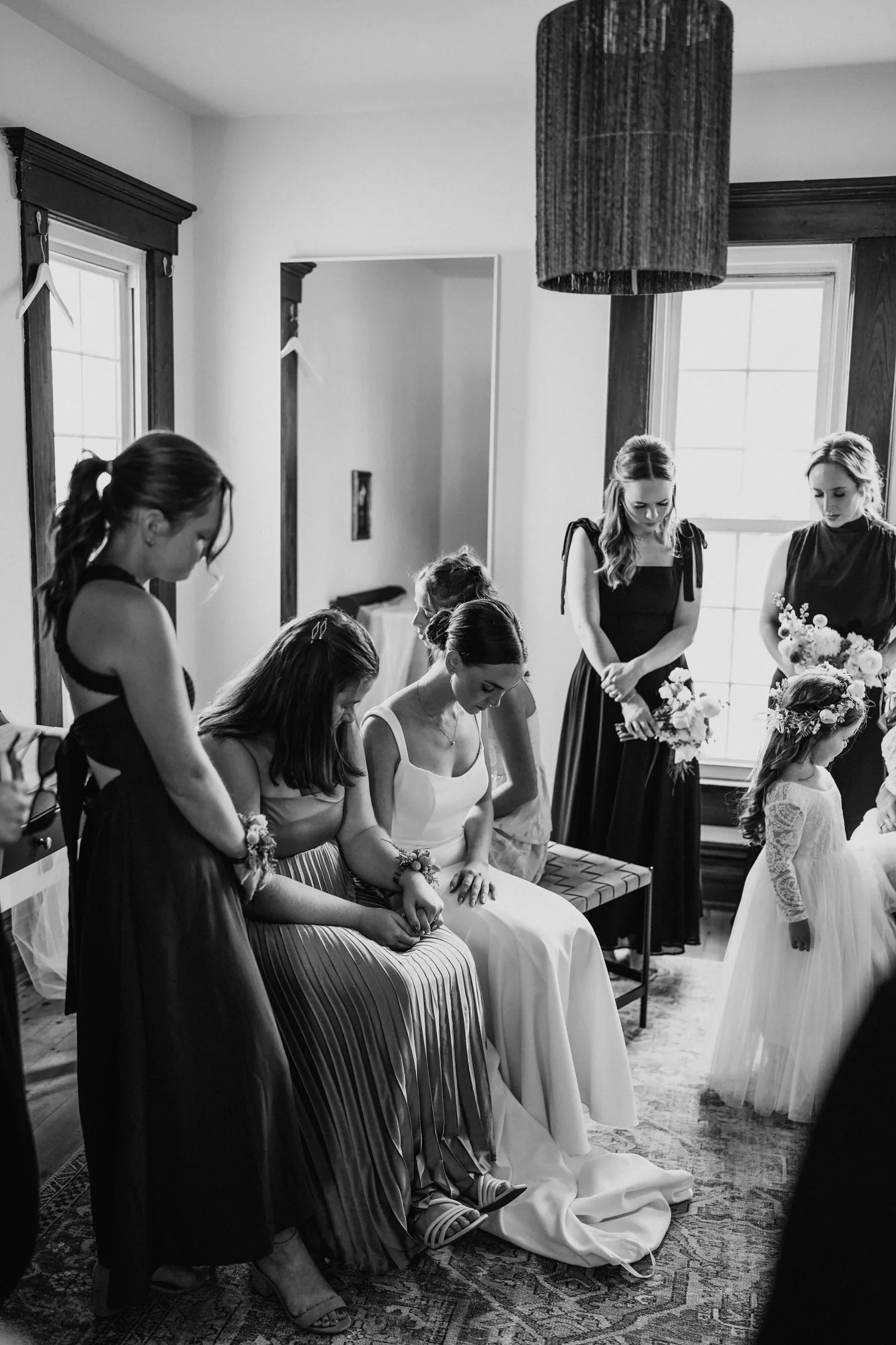 Group of women and a young girl gathered in a room, possibly preparing for a wedding, with some women in dresses and the girl in a bridal gown, all wearing corsages and bouquets.