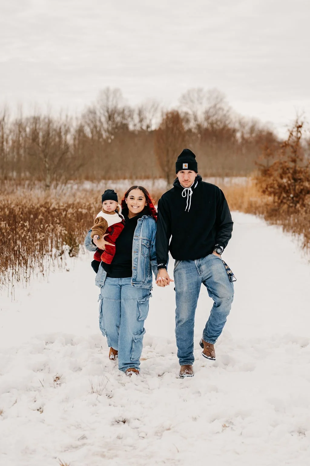 A family of three walking in the snow on a winter day, holding hands, with trees and a cloudy sky in the background.