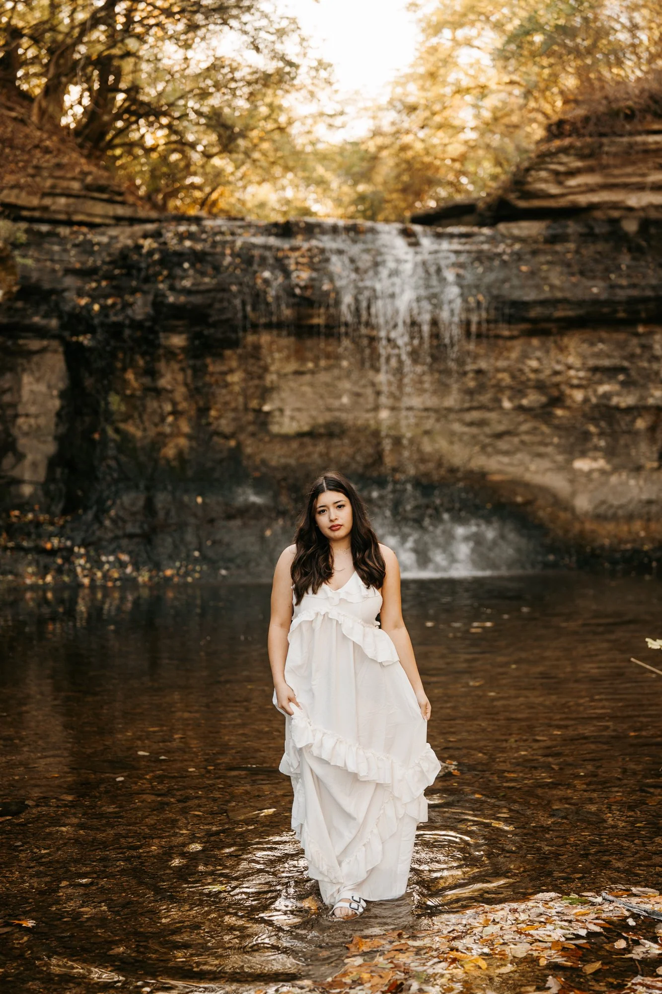 A young woman in a white dress standing in shallow water in front of a waterfall surrounded by autumn trees.
