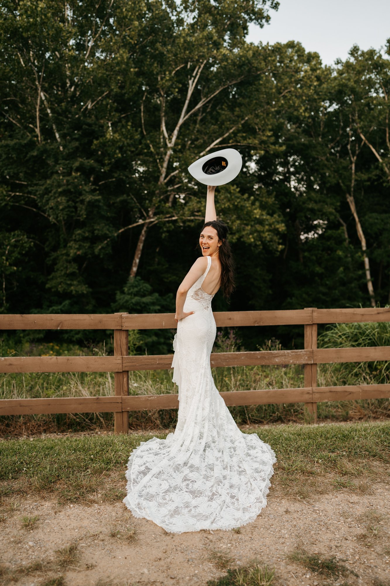 A woman in a white wedding dress holding a white wide-brimmed hat, smiling and looking back, outdoors near a wooden fence with lush green trees in the background.