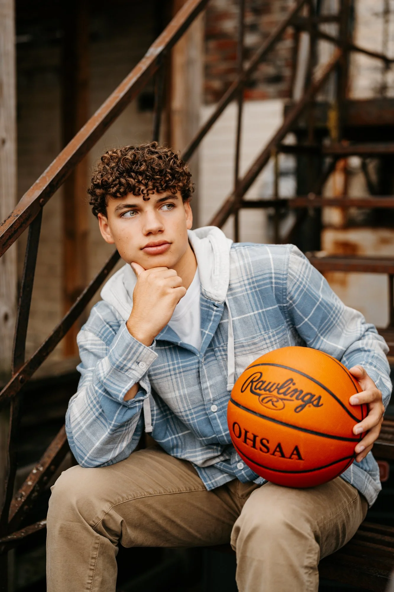 A young man with curly hair sitting on outdoor stairs, holding a basketball, and resting his chin on his hand while looking to the side.