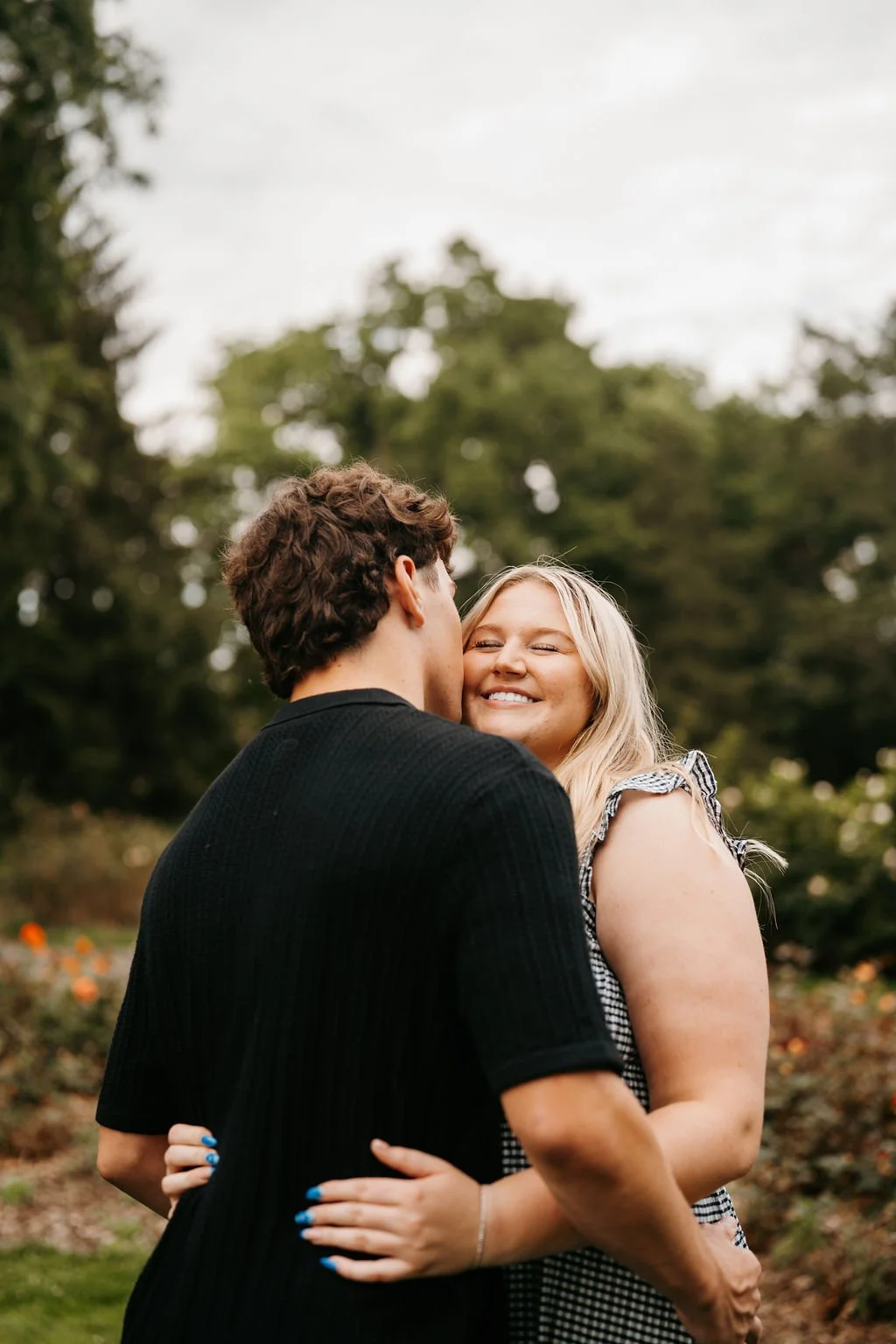 A man and woman embrace outdoors, with the man kissing the woman's cheek, and the woman smiling happily. They are surrounded by trees and foliage.