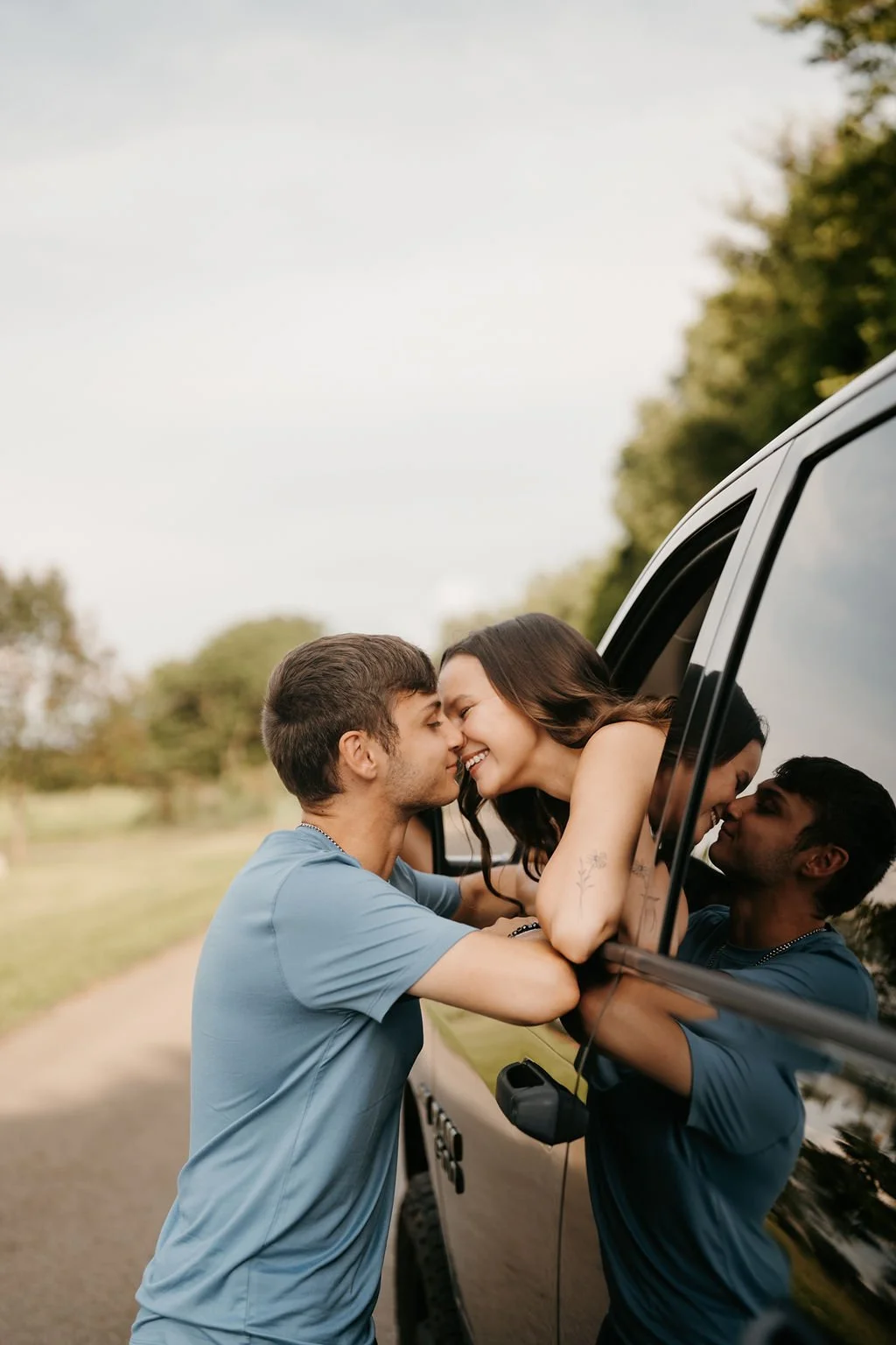 A couple leaning out of a vehicle window, touching foreheads and smiling at each other, with another person visible in the rearview mirror.