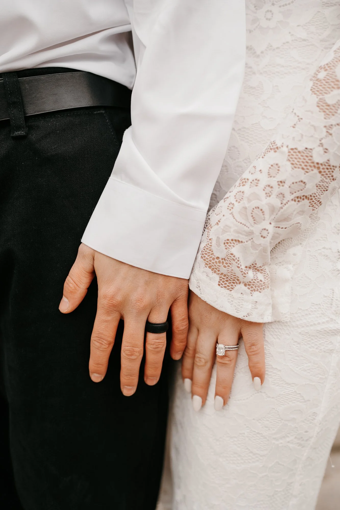 Close-up of a couple's hands with wedding rings, showing the groom wearing a black ring and the bride with a diamond engagement ring and a wedding band, against wedding attire.