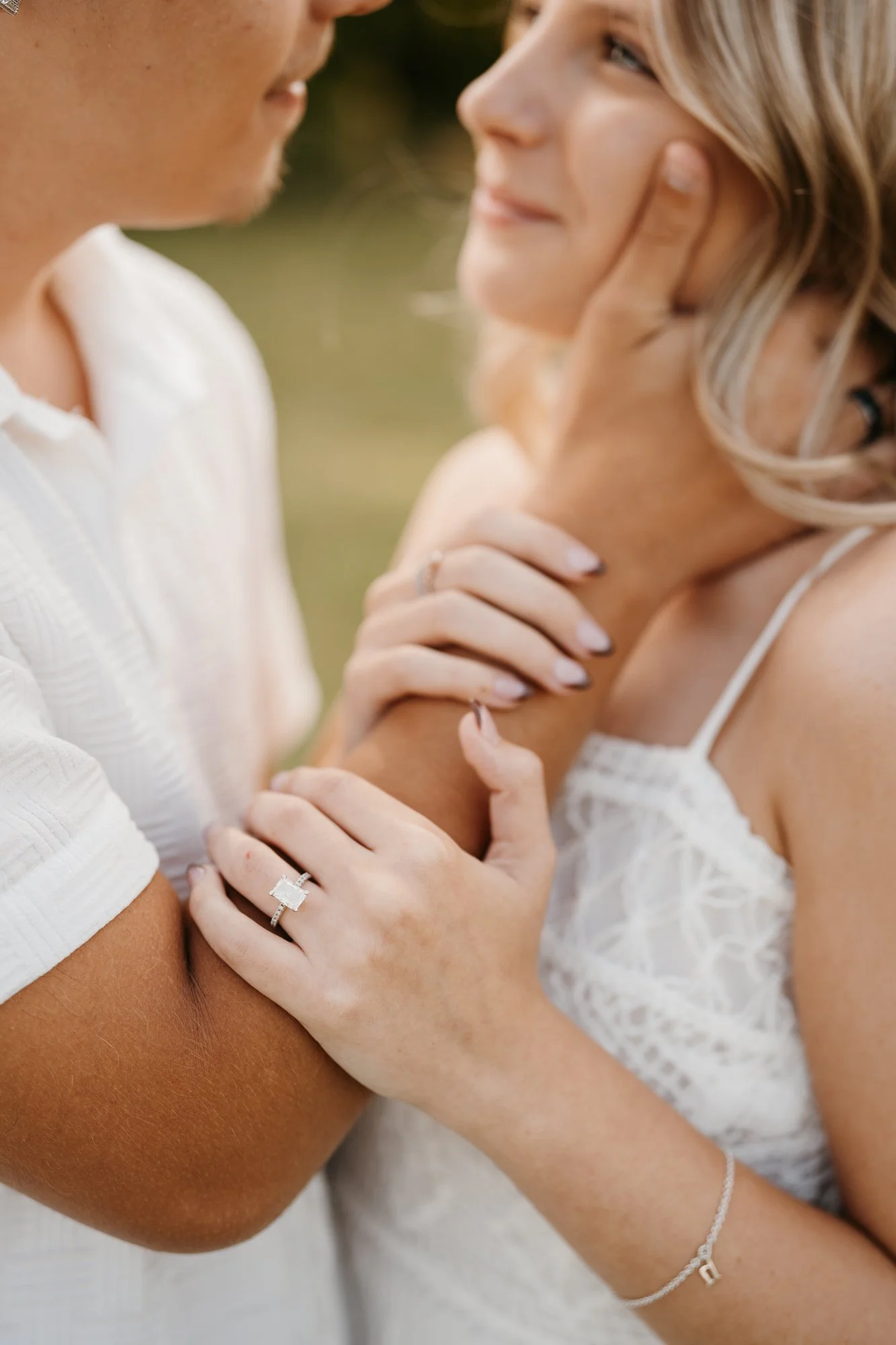 A woman and a man in an intimate moment, with the woman touching the man's face and holding her hand on his arm, wearing an engagement ring. They are outdoors with a blurred natural background.