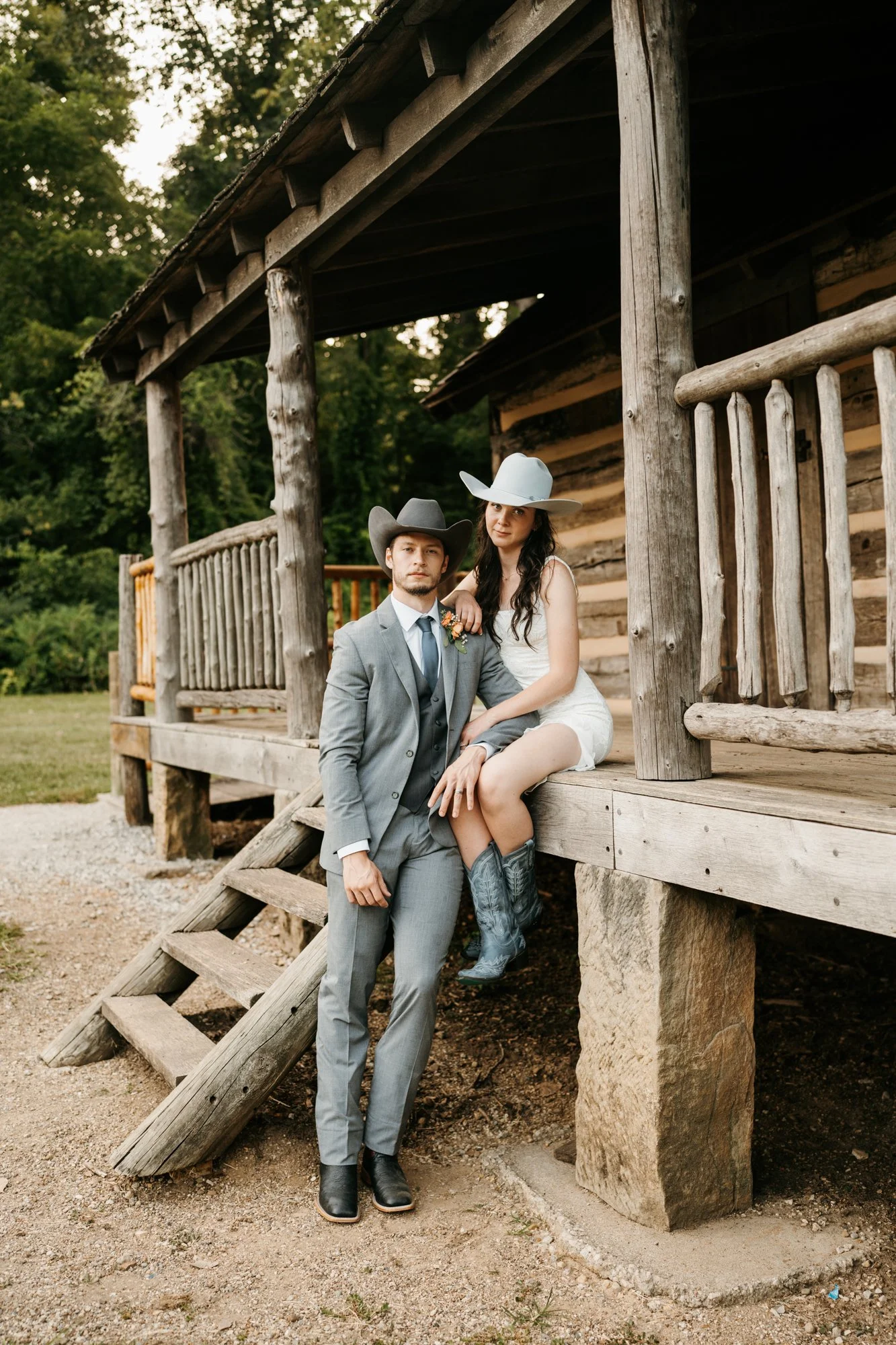 A man in a gray suit and cowboy hat sits on the steps of a wooden porch, with a woman in a white dress, cowboy hat, and cowboy boots sitting beside him. The scene is outdoors next to a rustic log cabin surrounded by trees.