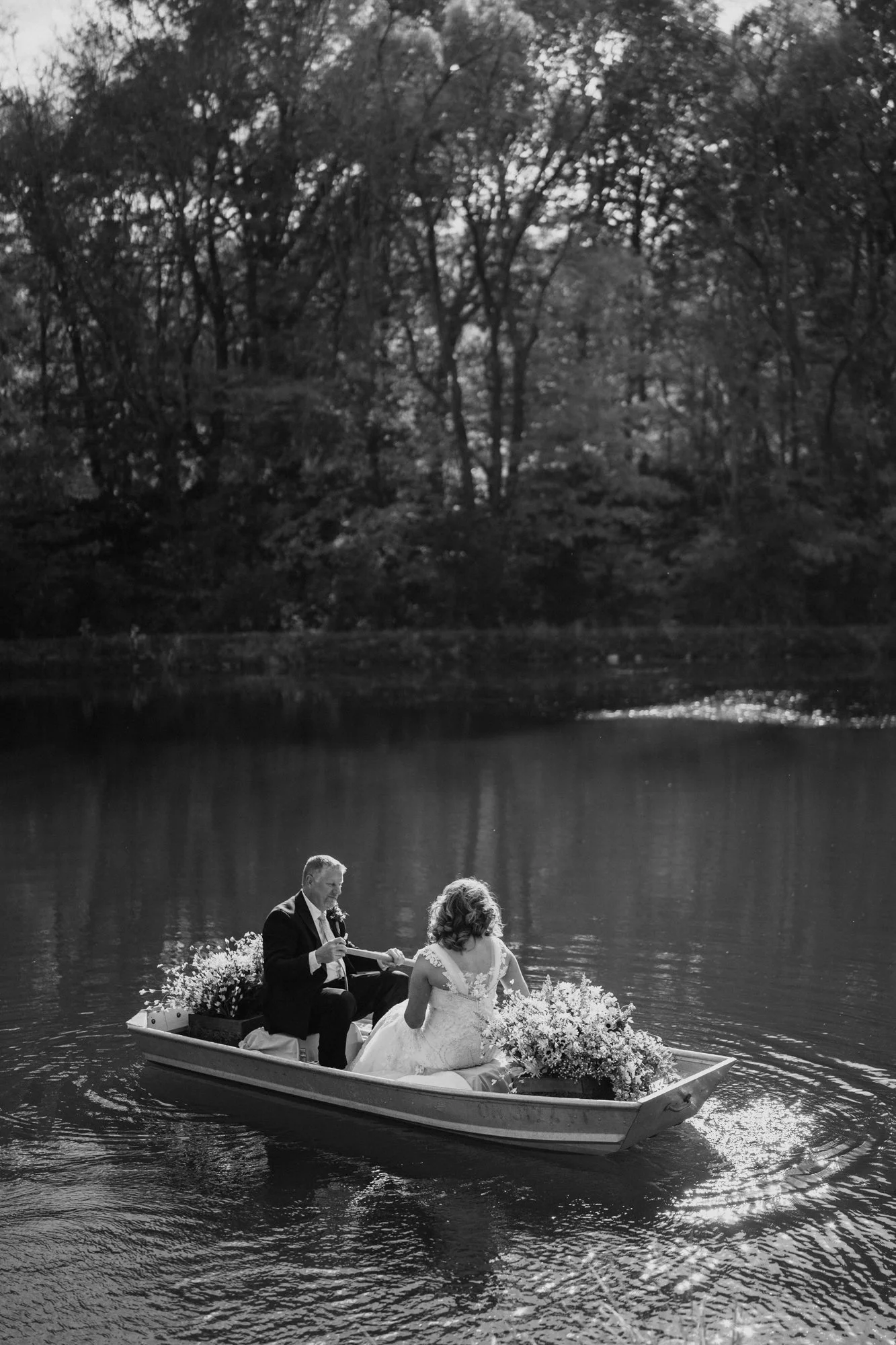 A wedding couple on a boat in a lake, with the groom in a suit and the bride in a wedding dress, surrounded by floral arrangements, with trees in the background.
