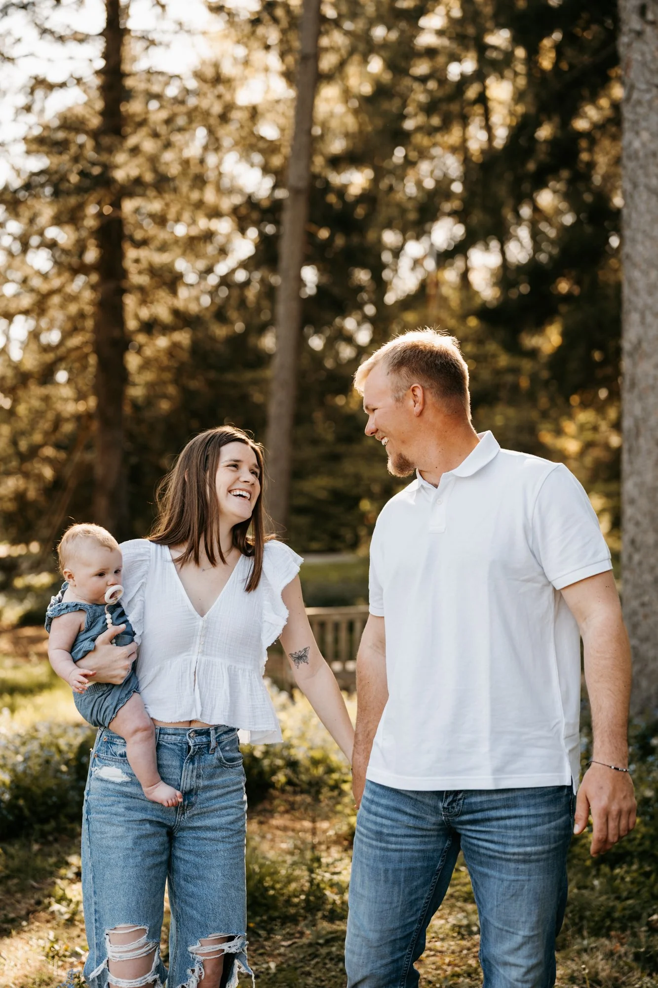 A family of three holding hands and smiling outdoors in a park during the golden hour, with trees in the background.