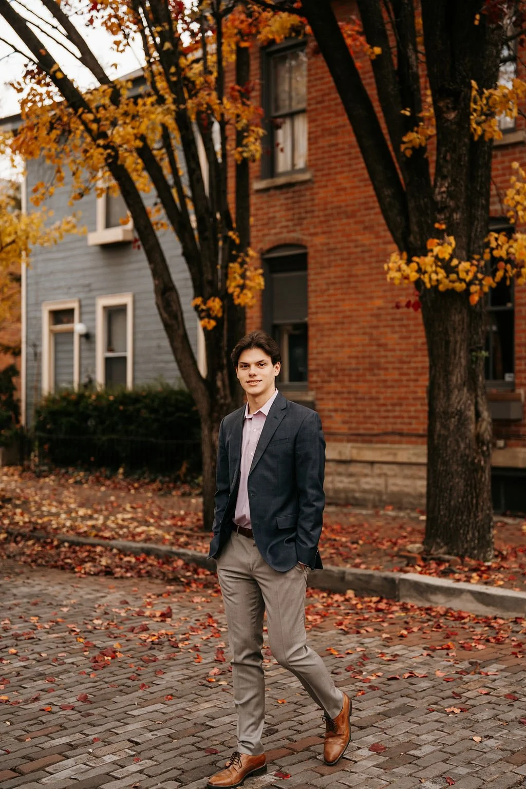 A young man in a gray blazer, pink shirt, gray pants, and brown shoes walking on a cobblestone street with fallen autumn leaves. Behind him are trees with yellow and orange leaves and brick and gray houses.