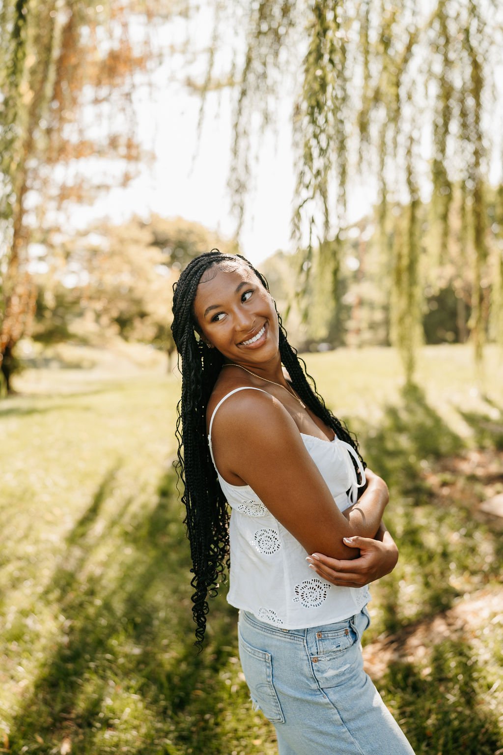 A young woman with braided hair and a white sleeveless top smiling outdoors in a park with green grass and trees.