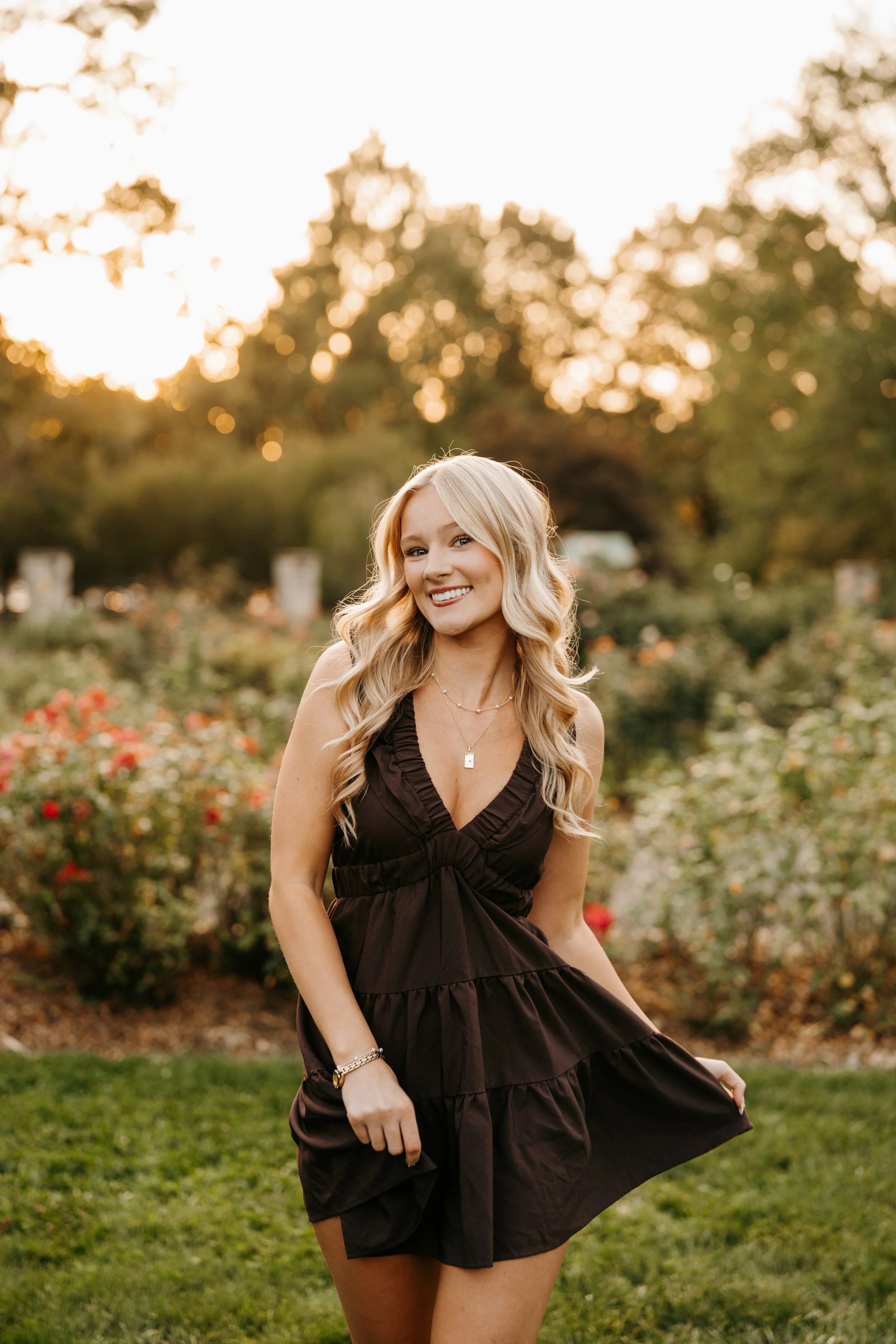 A young woman in a black dress smiling outdoors during sunset, standing in a garden with trees and flowers in the background.