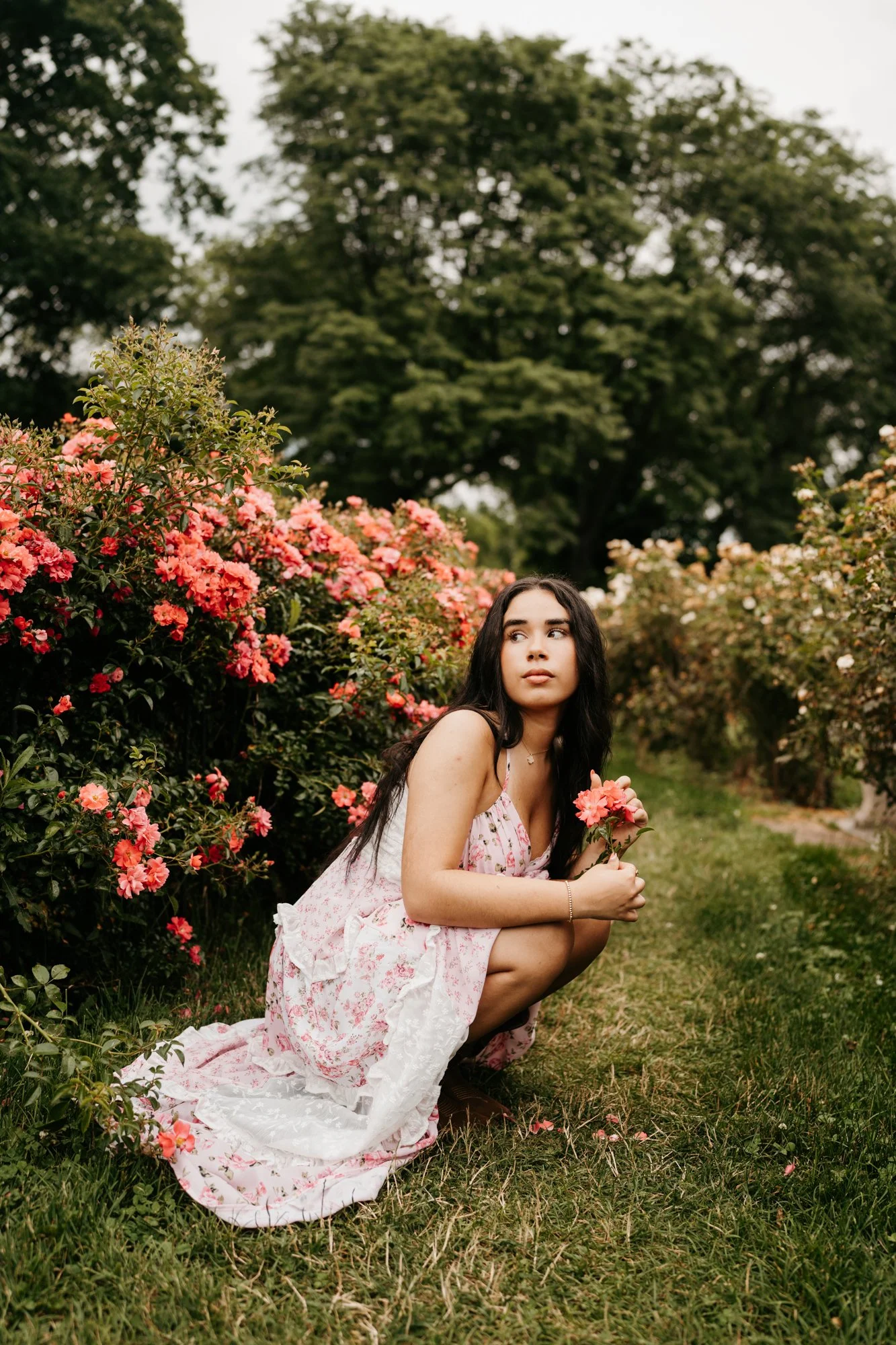 A young woman with long dark hair in a floral dress crouching in a garden surrounded by pink and white flowers, holding a small bouquet of flowers, with trees in the background.