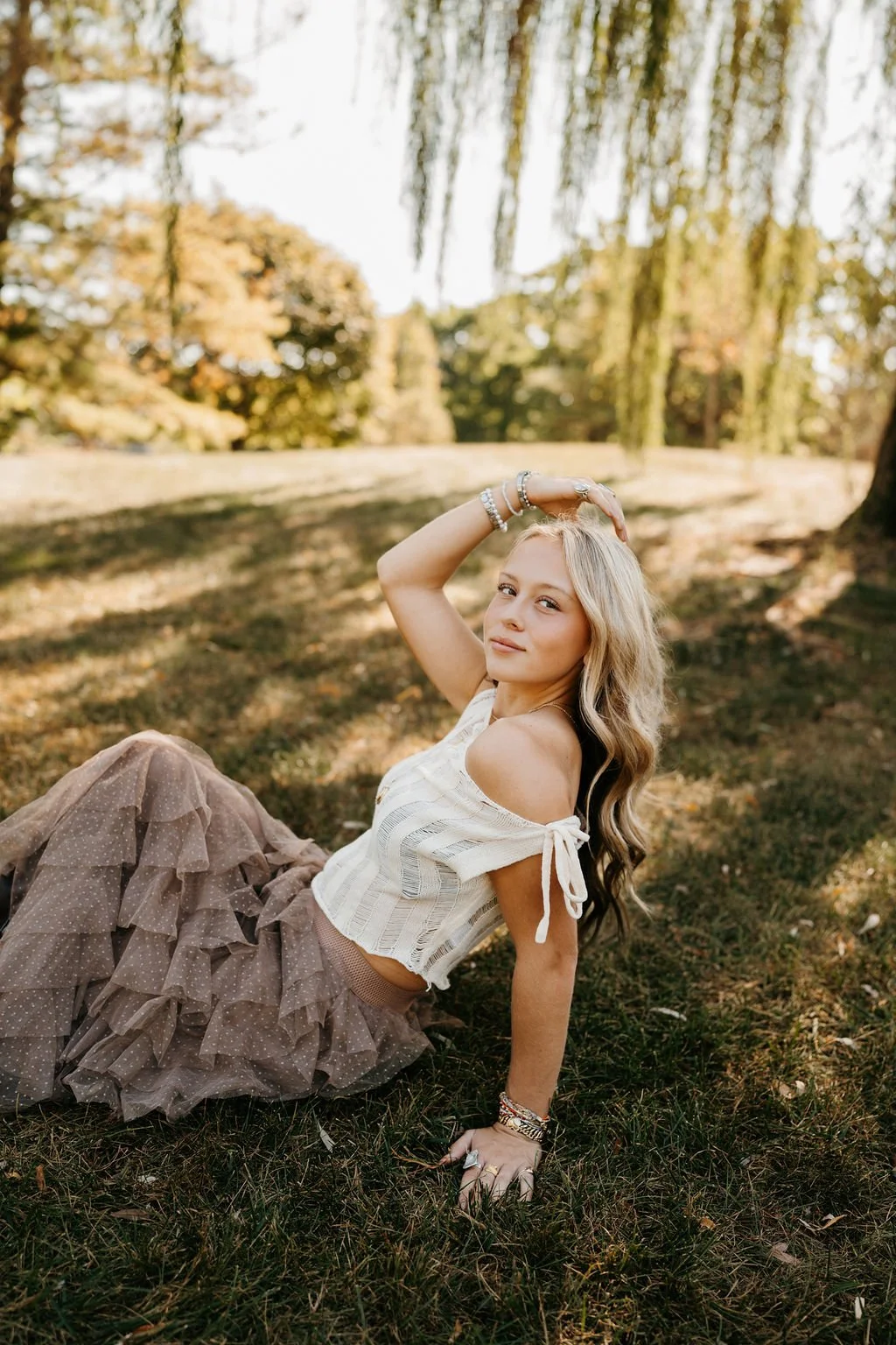 A young woman with long blond hair, wearing a white off-shoulder top and a brown layered tulle skirt, sitting on the grass in a park with trees and autumn foliage in the background.