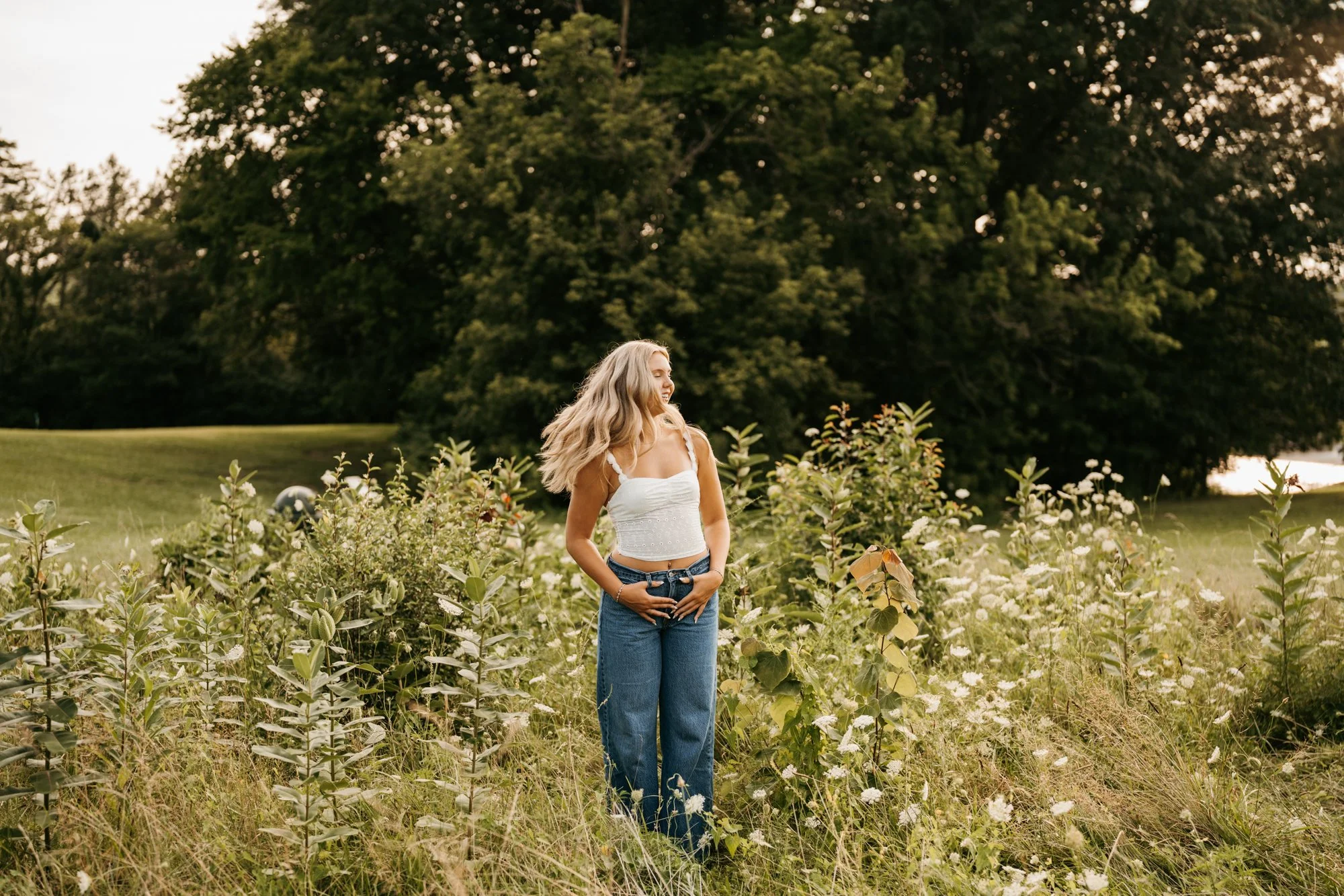 A young woman with long blonde hair standing in a grassy field with wildflowers, wearing a white crop top and blue jeans, smiling and looking away.
