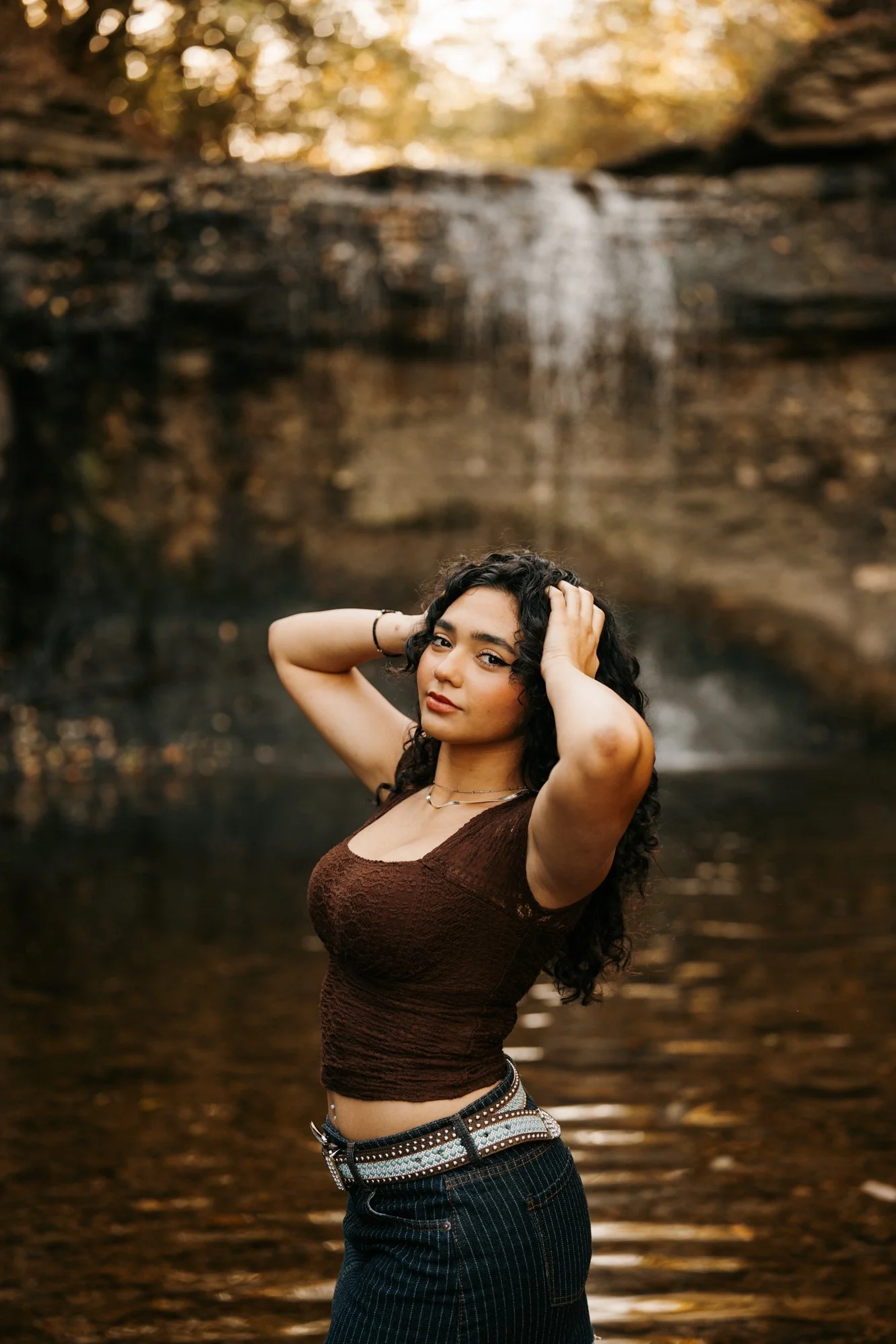 Young woman with curly hair posing in front of a waterfall in a natural setting during daytime.